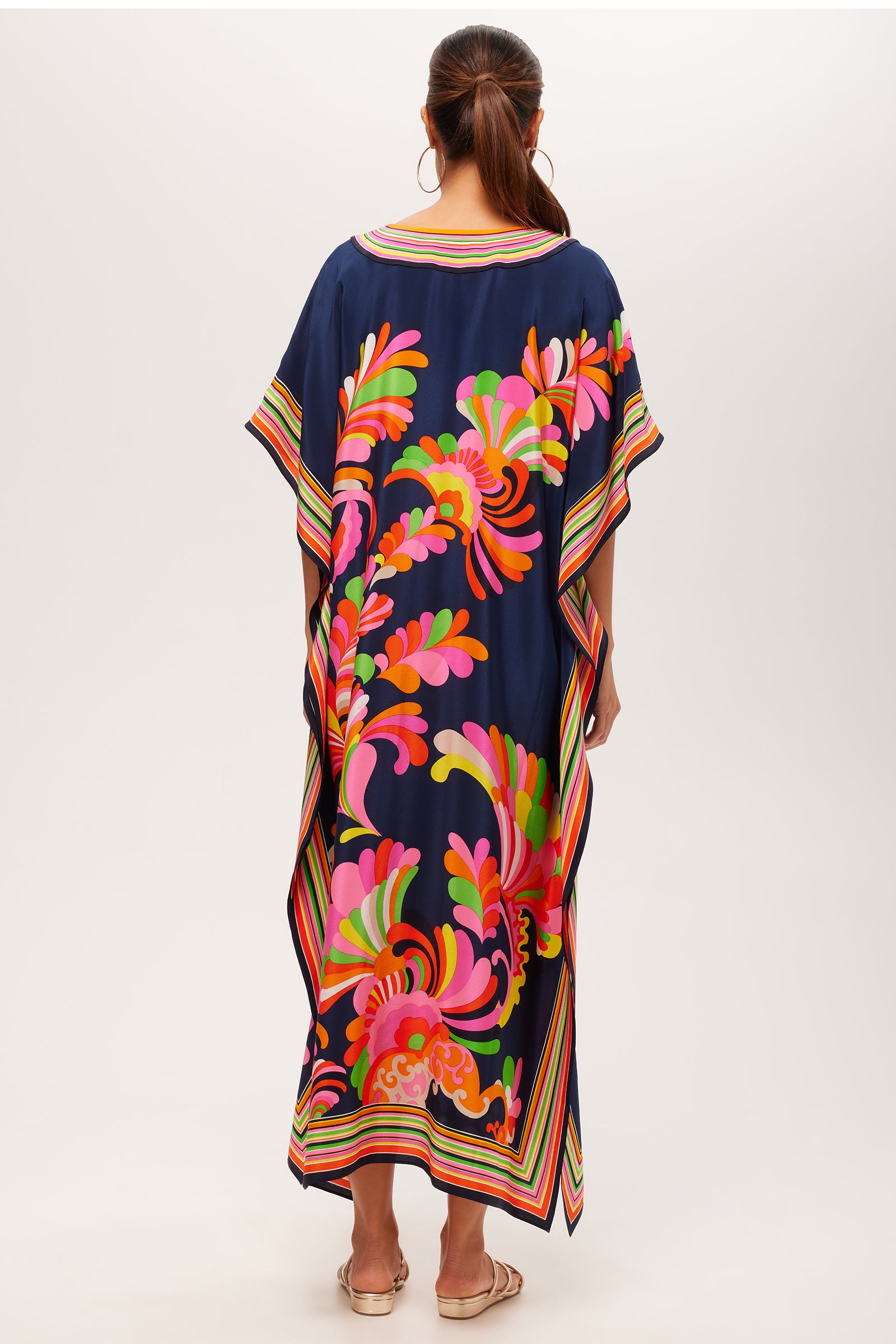 A woman with brown hair in a ponytail stands with her back to the camera, wearing the Theodora Maxi Caftan - Paradise Palms Silk, featuring bold pink, orange, and green floral and leaf patterns and multicolored striped borders.