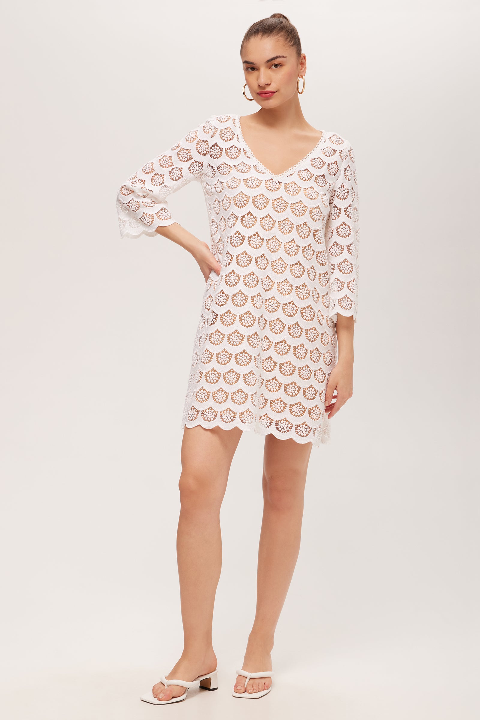 A woman poses with one hand on her hip, wearing the white Lita Crochet 3/4-Sleeve Dress featuring a knee-length cut and V-neck. Her hair is tied back, and she accessorizes with hoop earrings and white heeled sandals.