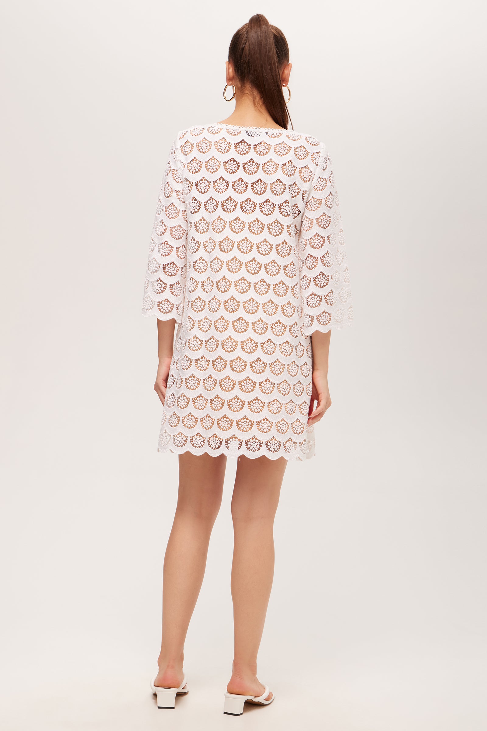 A woman with long brown hair in a ponytail wears the Lita Crochet 3/4-Sleeve Dress—white, knee-length—paired with white heeled sandals and hoop earrings. She stands facing away against a plain light gray background.