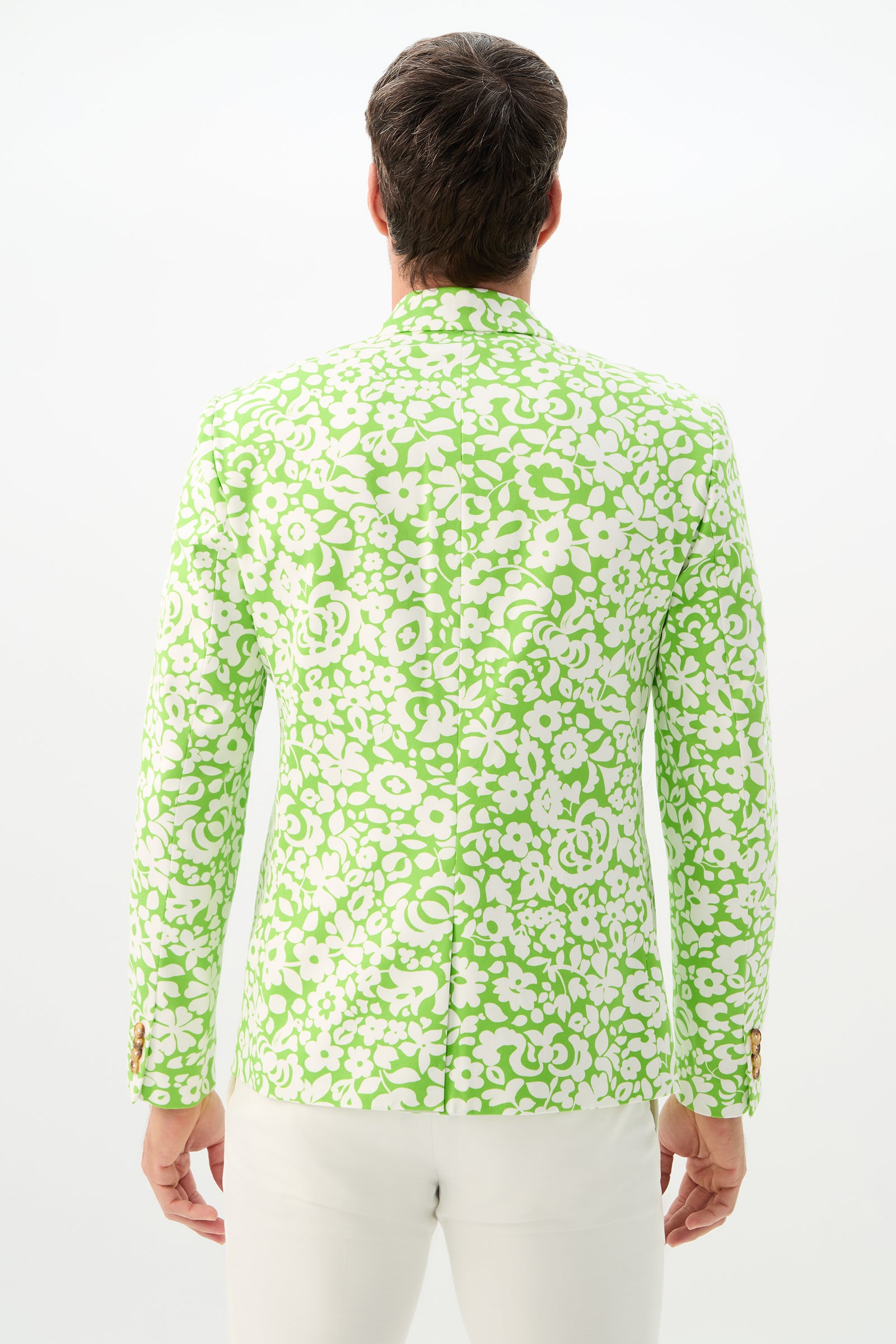 A person stands facing away, wearing the Thurston Blazer - Poolside Gossip, featuring a green and white floral pattern, paired with white pants against a plain white background.