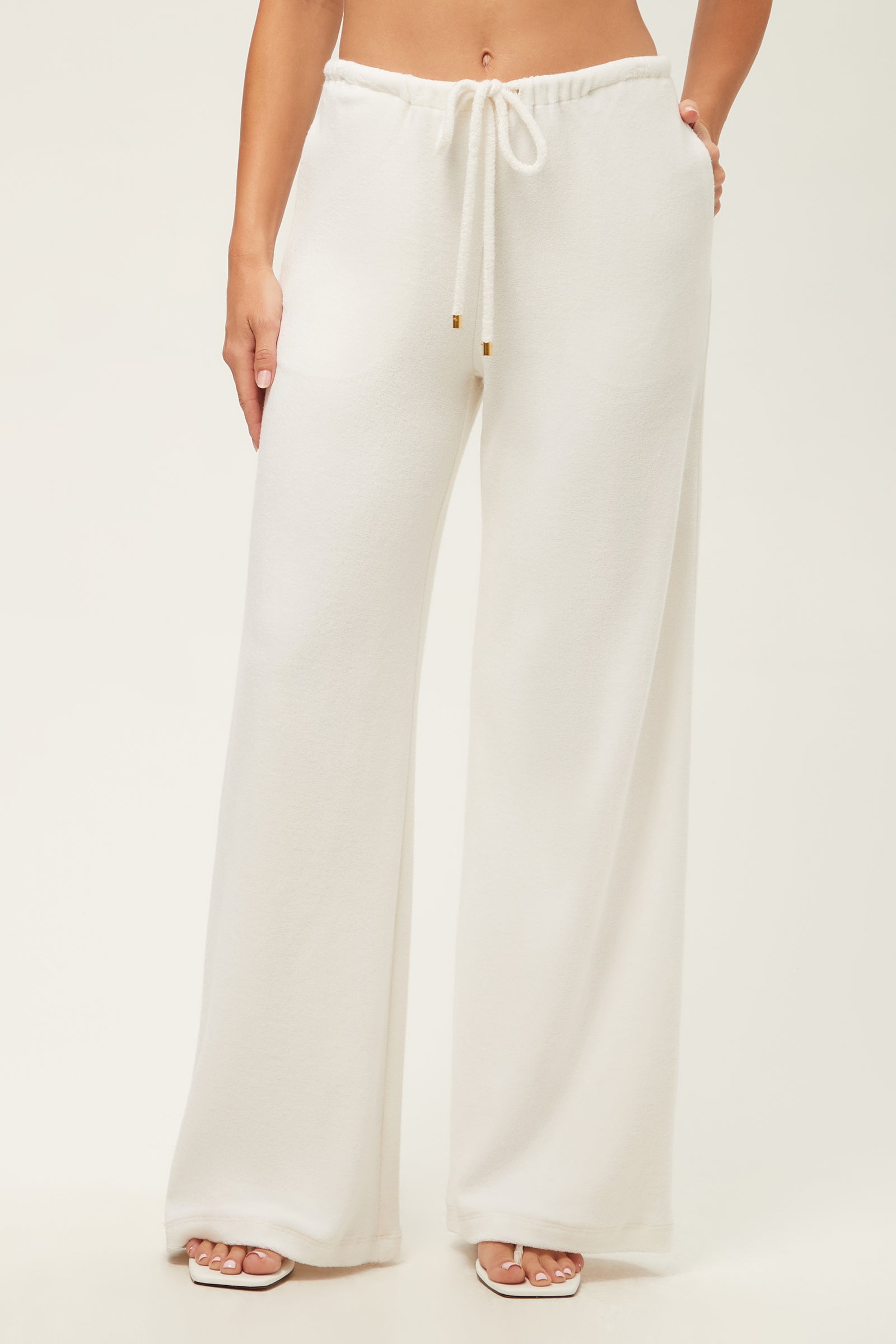 A woman, barefoot against a plain light background, wears the Linsk Wide-Leg Pant - Poolside Terry in white; the high-waisted pants feature a wide leg and drawstring waist. Her top and face are not shown.