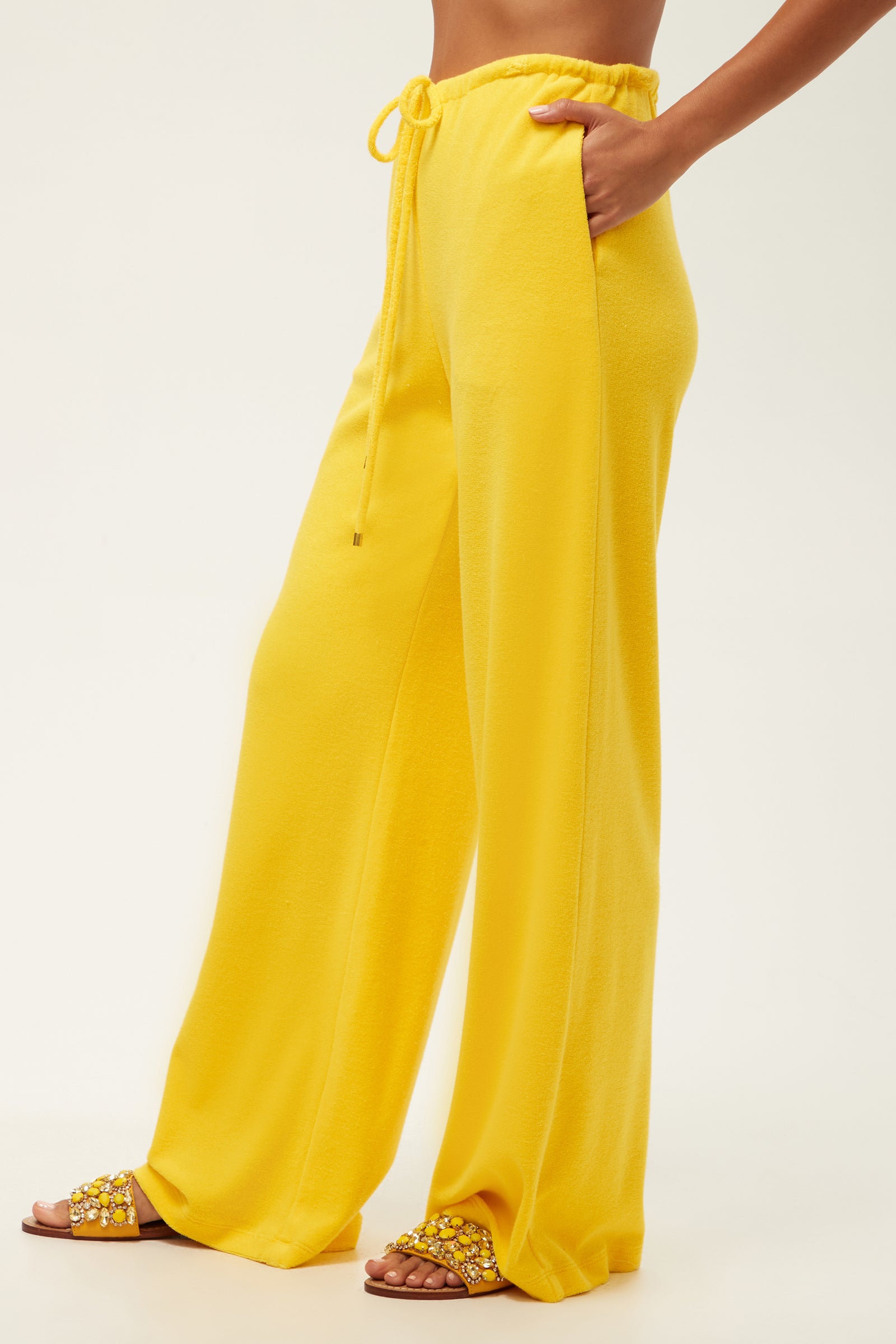 A person wears the Linsk Wide-Leg Pant - Poolside Terry in bright yellow with a drawstring waist and matching embellished sandals, standing against a plain light background.