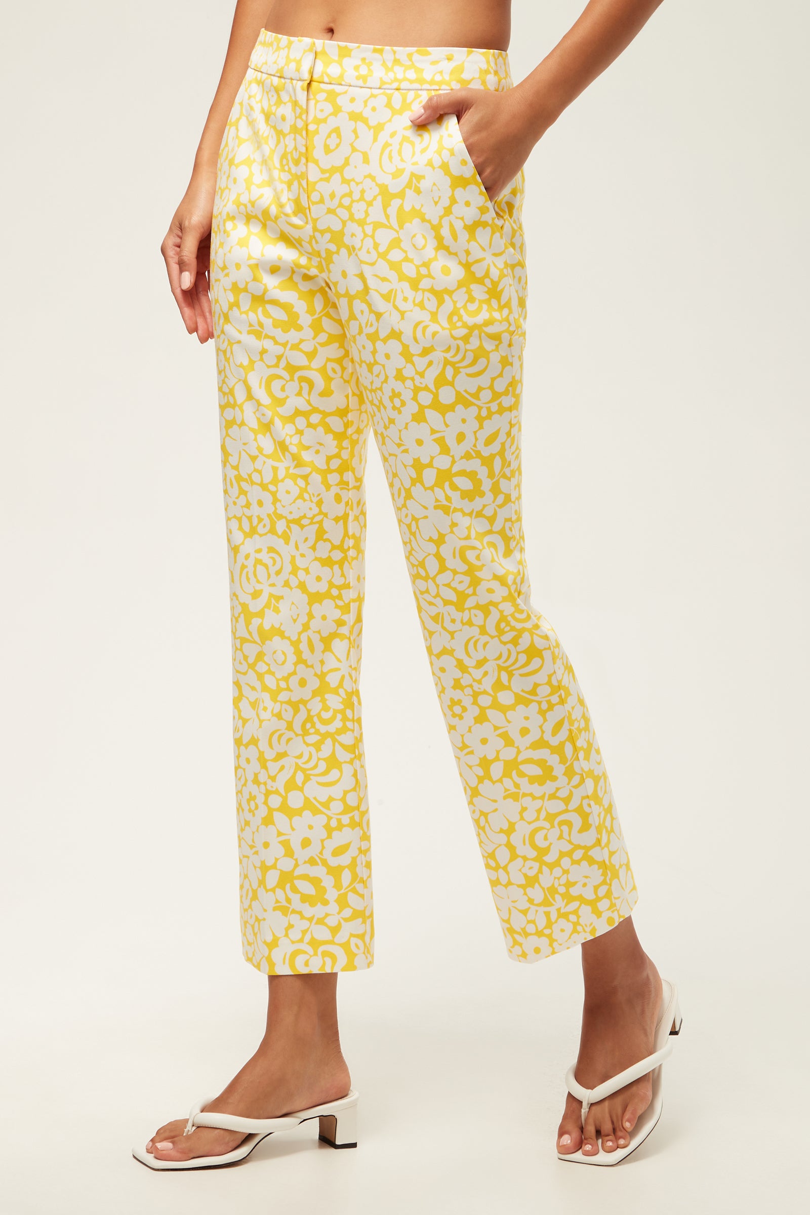 A woman models the Lulu Cropped Pant - Poolside Gossip, featuring a yellow and white floral print in a straight-leg, cropped fit with front pockets. She pairs them with white heeled sandals; only her torso and legs show against a plain background.