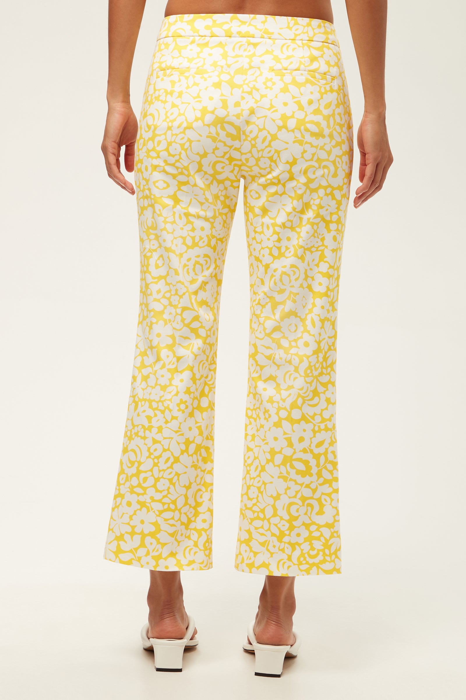 A person is seen from behind wearing the Lulu Cropped Pant - Poolside Gossip, featuring a yellow and white floral pattern with a slight flare, paired with white open-toe heeled sandals against a plain light background.