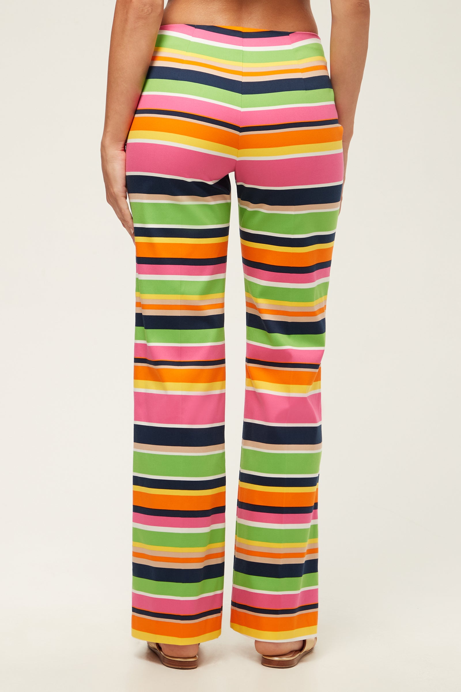 A person stands facing away in the Rudi Straight-Leg Pant – Splash Stripe, featuring high-waisted, flared legs with bold pink, green, orange, yellow, black, and white horizontal stripes. The background is plain and light-colored.