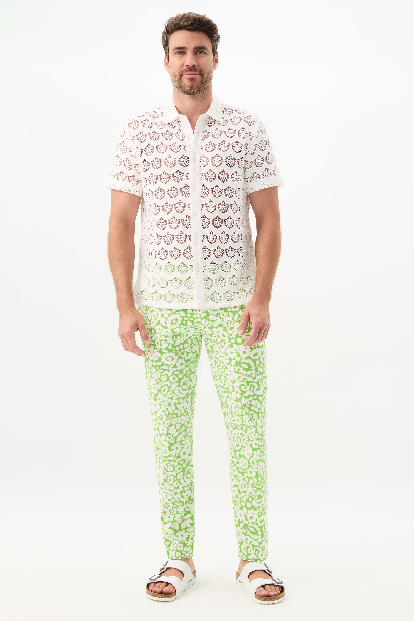 A man with short brown hair and a beard, smiling slightly, stands against a white background wearing the Alex Slim Trouser - Poolside Gossip, paired with a sheer white patterned shirt and white sandals.