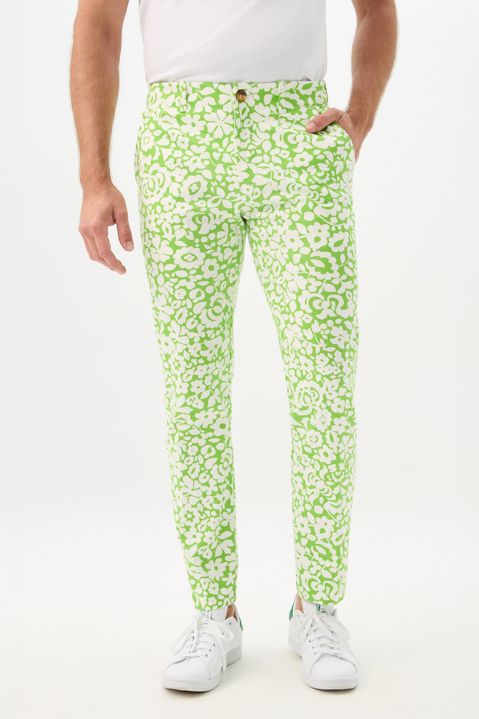 A person wears the Alex Slim Trouser – Poolside Gossip in Serene Green with a white t-shirt and sneakers, standing with one hand in their pocket against a plain white background.