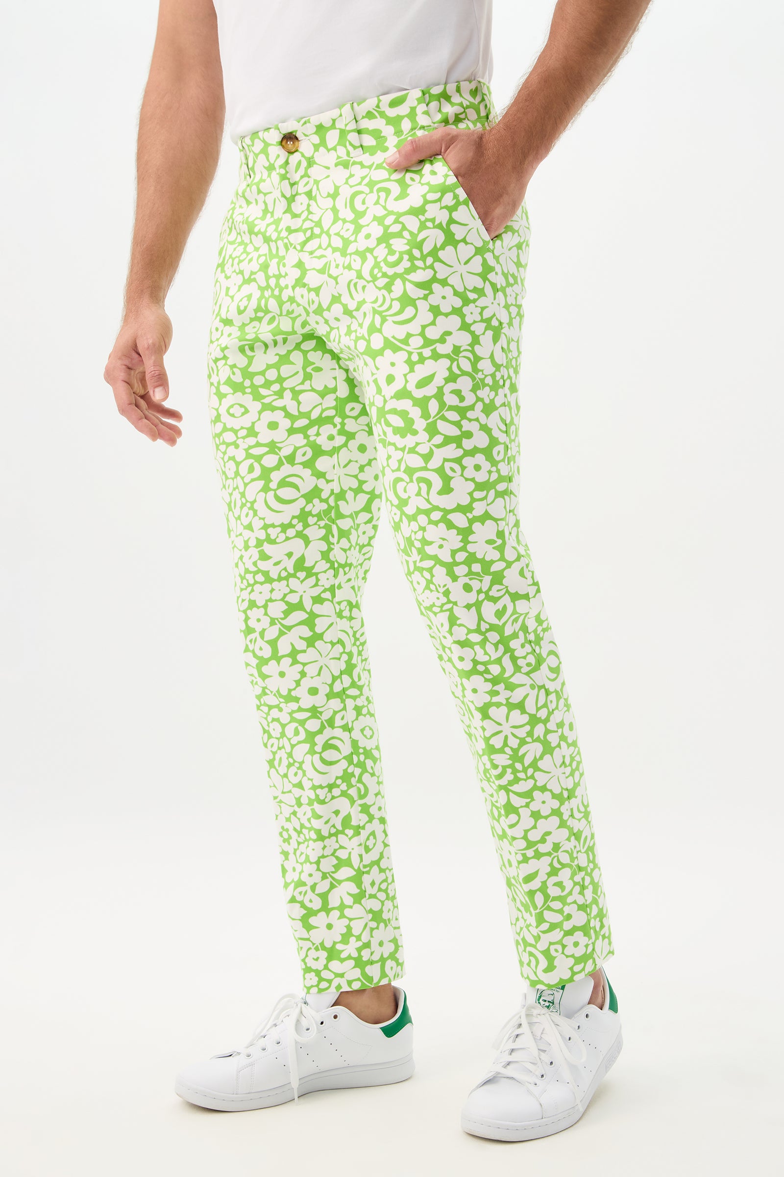 A person wearing the Alex Slim Trouser - Poolside Gossip, featuring a green and white floral pattern, stands with one hand in their pocket. Only their lower body is visible against a plain white background. White sneakers complete the look.