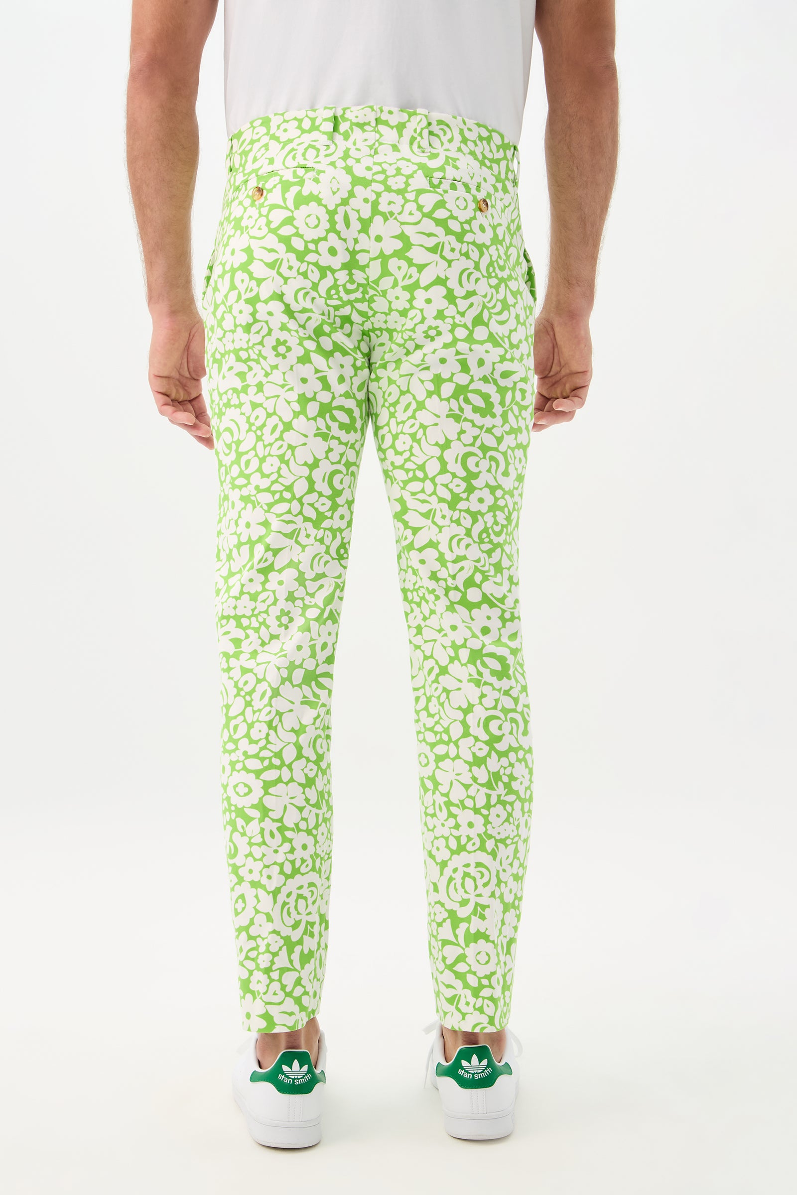 A person modeling the Alex Slim Trouser - Poolside Gossip, featuring a bright green floral print, paired with a white t-shirt and green and white Adidas sneakers, stands with their back to a plain white background.