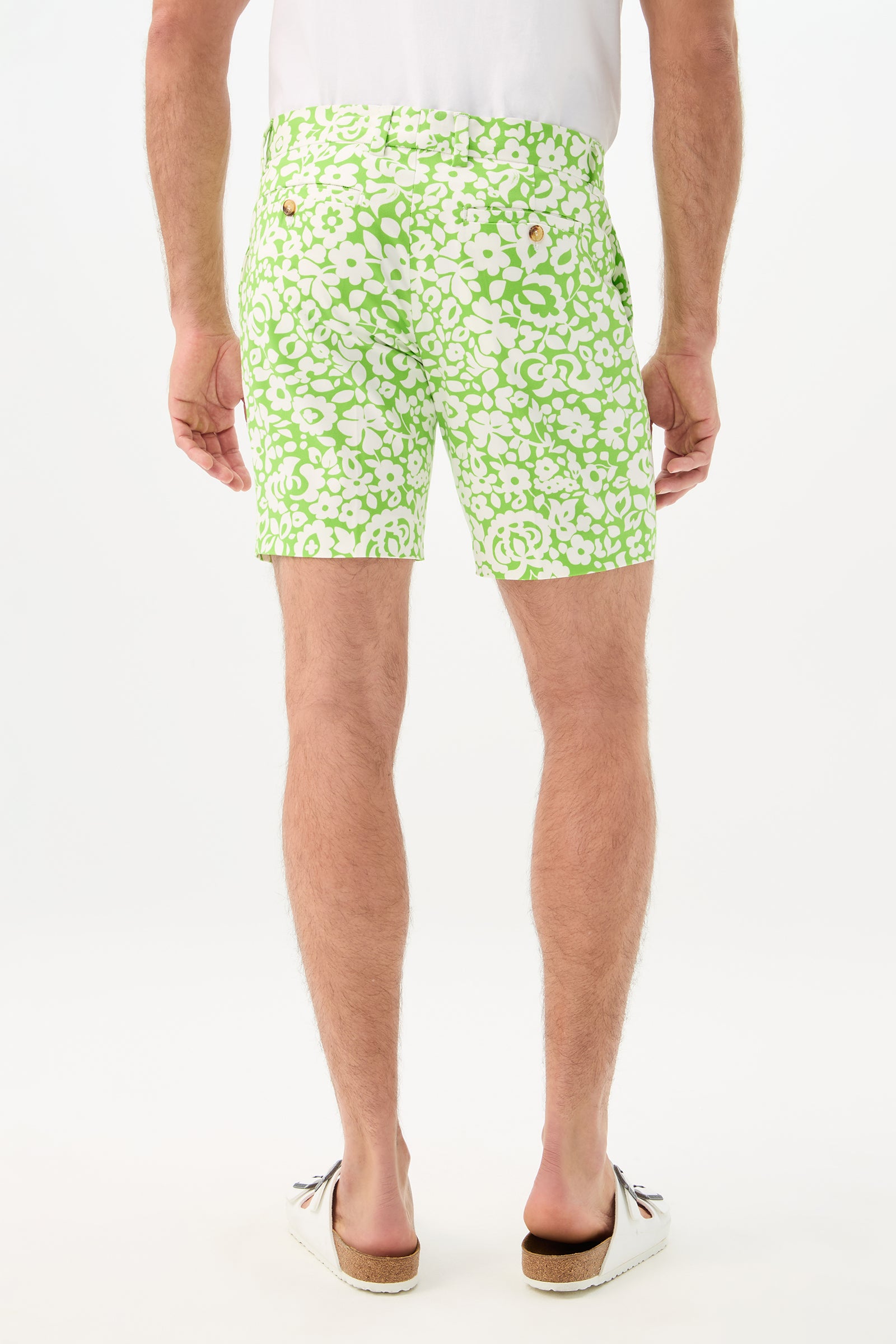 A man faces away, wearing the Lawrence Short - Poolside Gossip with a green and white floral print, paired with a white shirt and white slip-on shoes with brown soles, set against a plain white background.