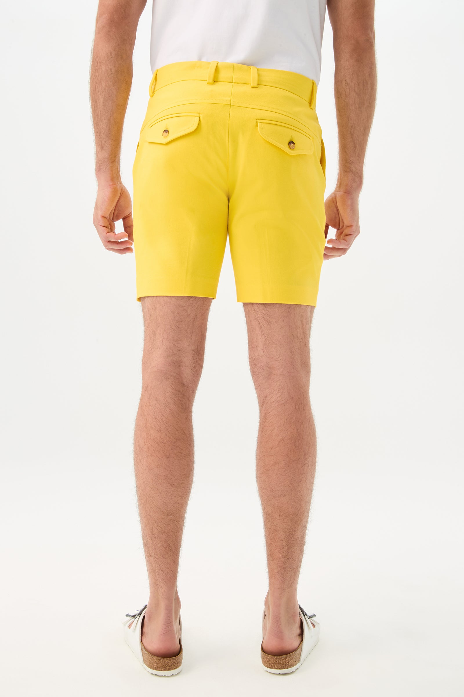 A man faces away from the camera, wearing a white t-shirt, Cyrus Short - Sunset Stretch in bright yellow with belted waist and buttoned back pockets, and striped slip-on shoes, set against a plain white background.