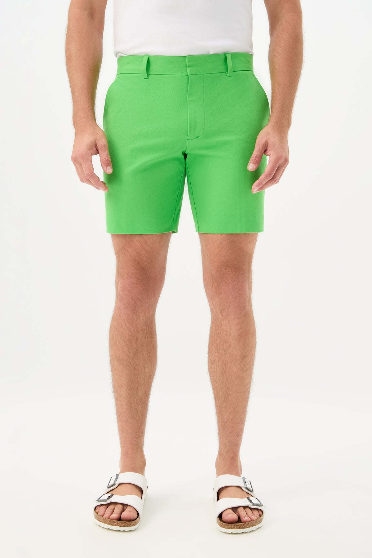 A person is shown from the waist down, wearing the Cyrus Short - Cotton Bi-Stretch in vert with a white shirt and white sandals, standing against a plain white background.