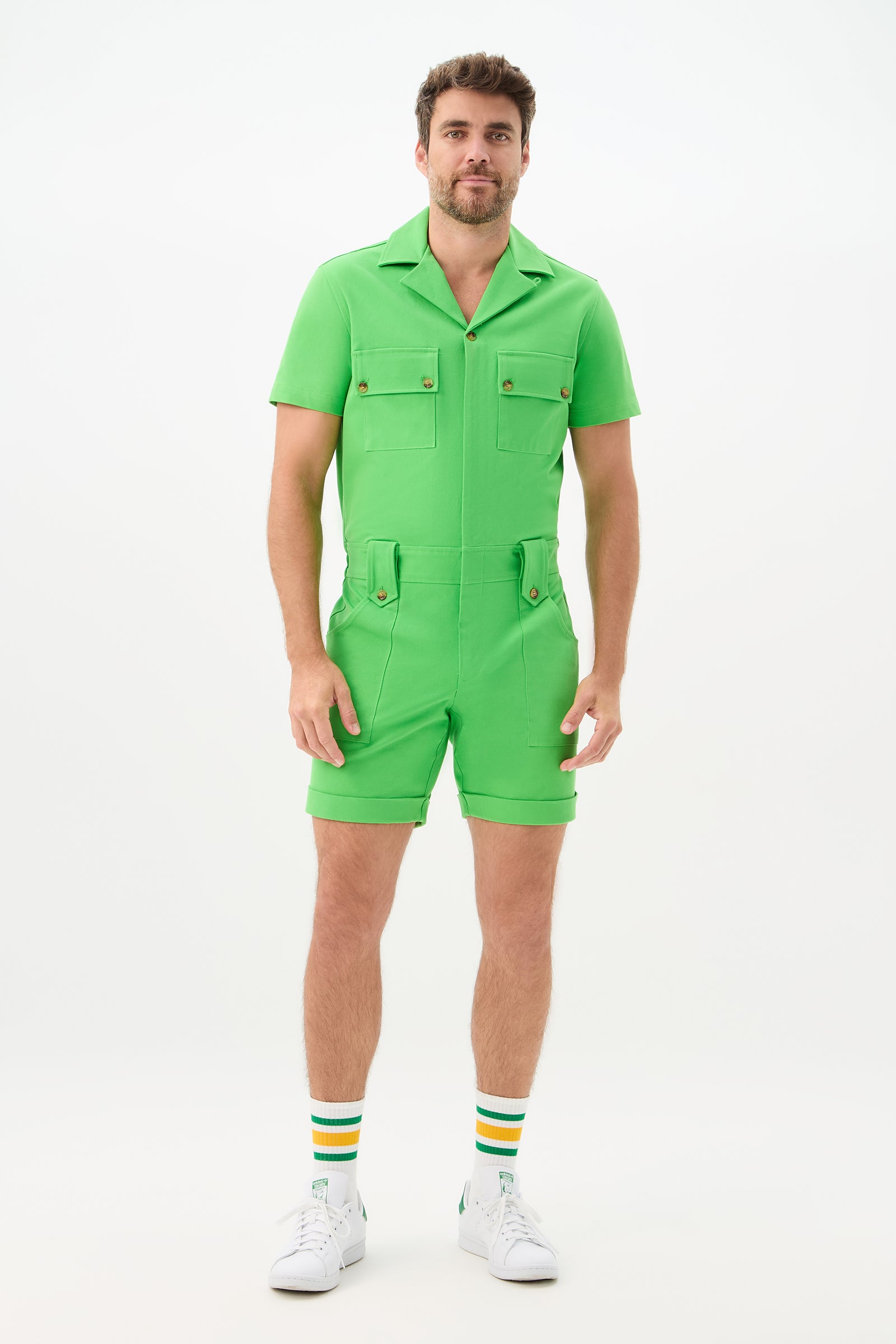 A man faces forward on a plain white background, wearing the Jaden 2 Short Jumpsuit - Cotton Bi-Stretch in bright green, paired with white sneakers and white socks featuring yellow and green stripes.