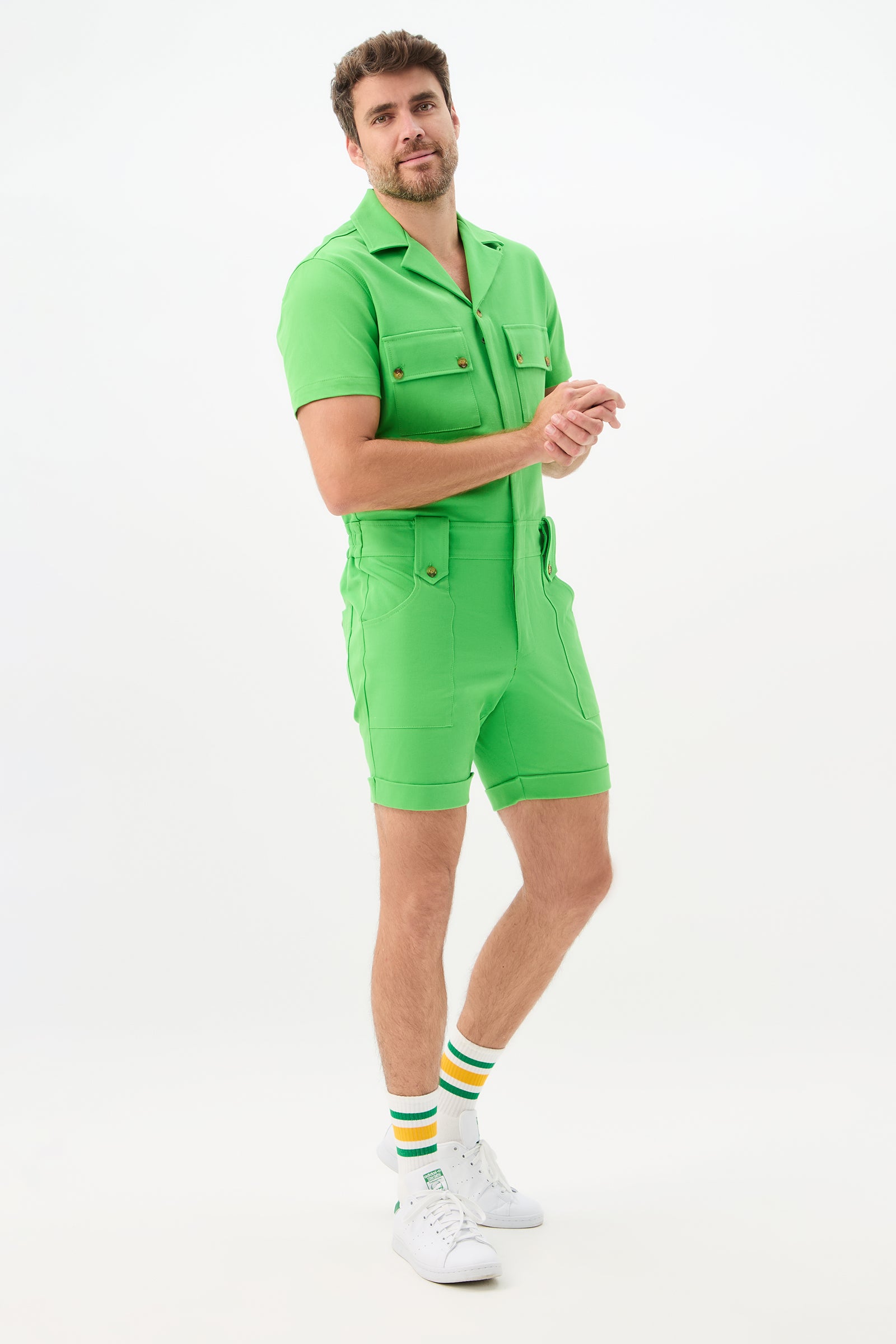 A man with a light beard smiles and looks to the side, wearing the bright green Jaden 2 Short Jumpsuit - Cotton Bi-Stretch, paired with white sneakers and white socks featuring green and yellow stripes, against a white background.