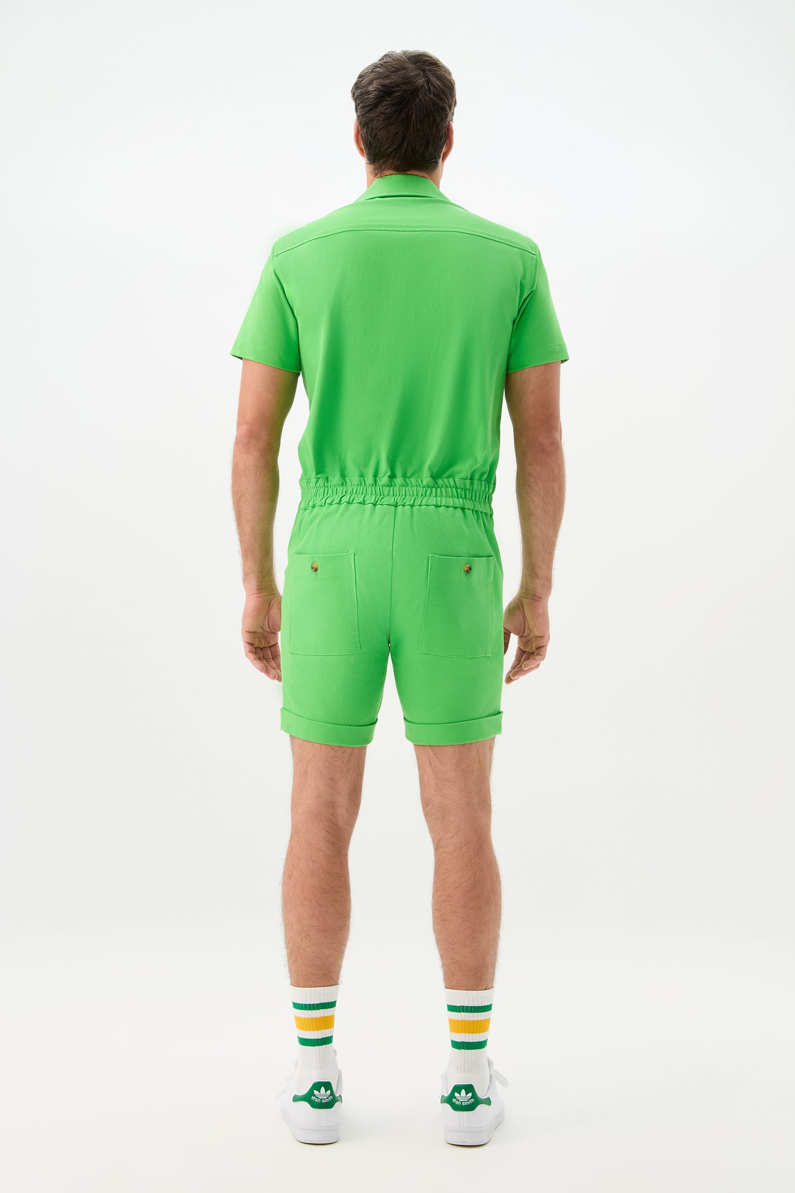A man faces away from the camera, wearing the Jaden 2 Short Jumpsuit - Cotton Bi-Stretch in bright green, paired with white socks featuring yellow and green stripes and white sneakers with green accents, set against a plain white background.