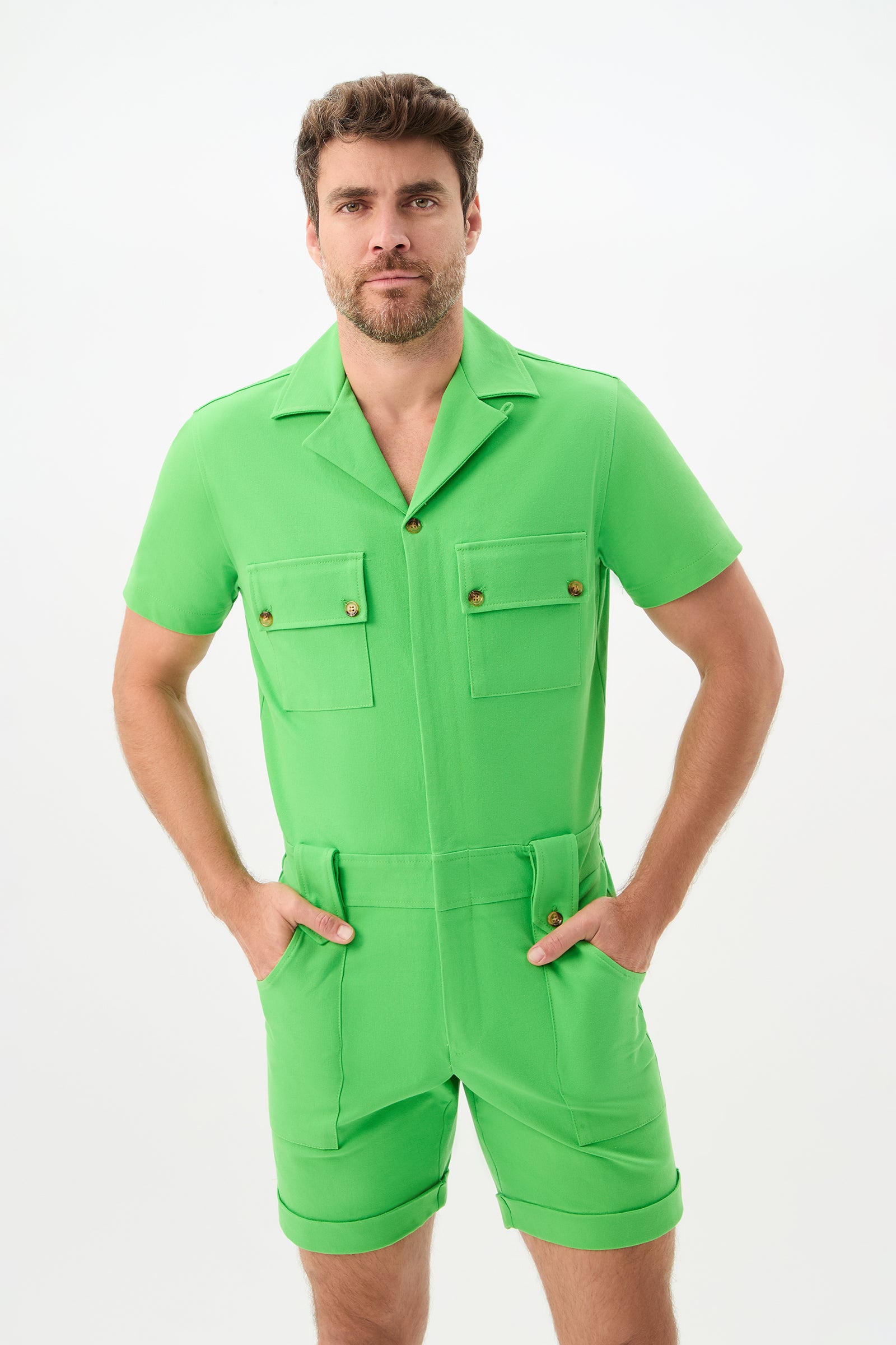 A man with short brown hair and a beard wears the Jaden 2 Short Jumpsuit in Vert, featuring pockets and buttons, standing against a plain white background with his hands in the front pockets.