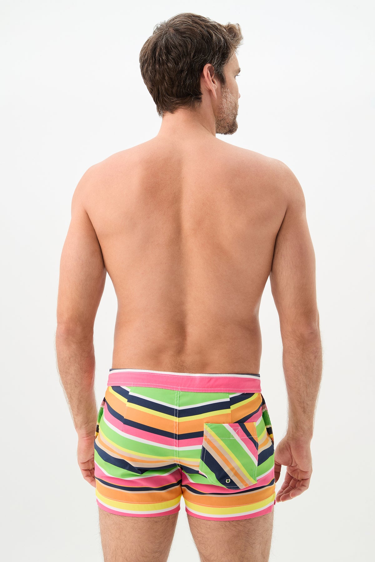 A man faces away from the camera, modeling the Chico Swim Trunk - Splash Stripe, which features colorful diagonal stripes in pink, yellow, green, orange, and black against a plain white background.
