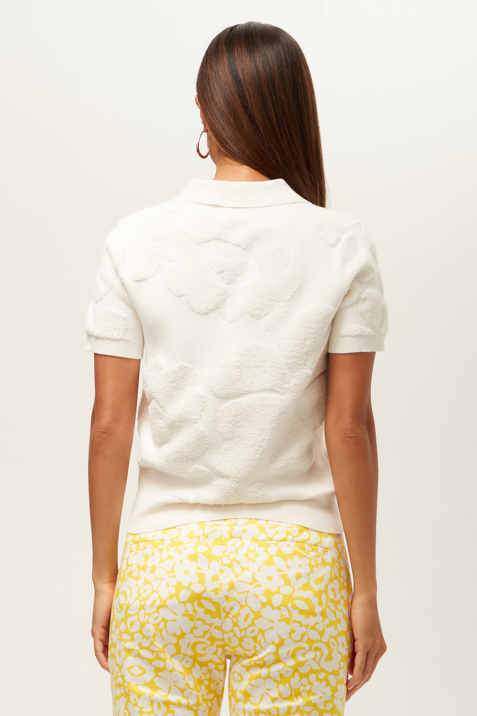 A woman with straight brown hair wears the Guiness Short-Sleeve Polo Top - Linsk Terry in white, paired with yellow floral pants and gold hoop earrings. She is facing away against a plain, light background.