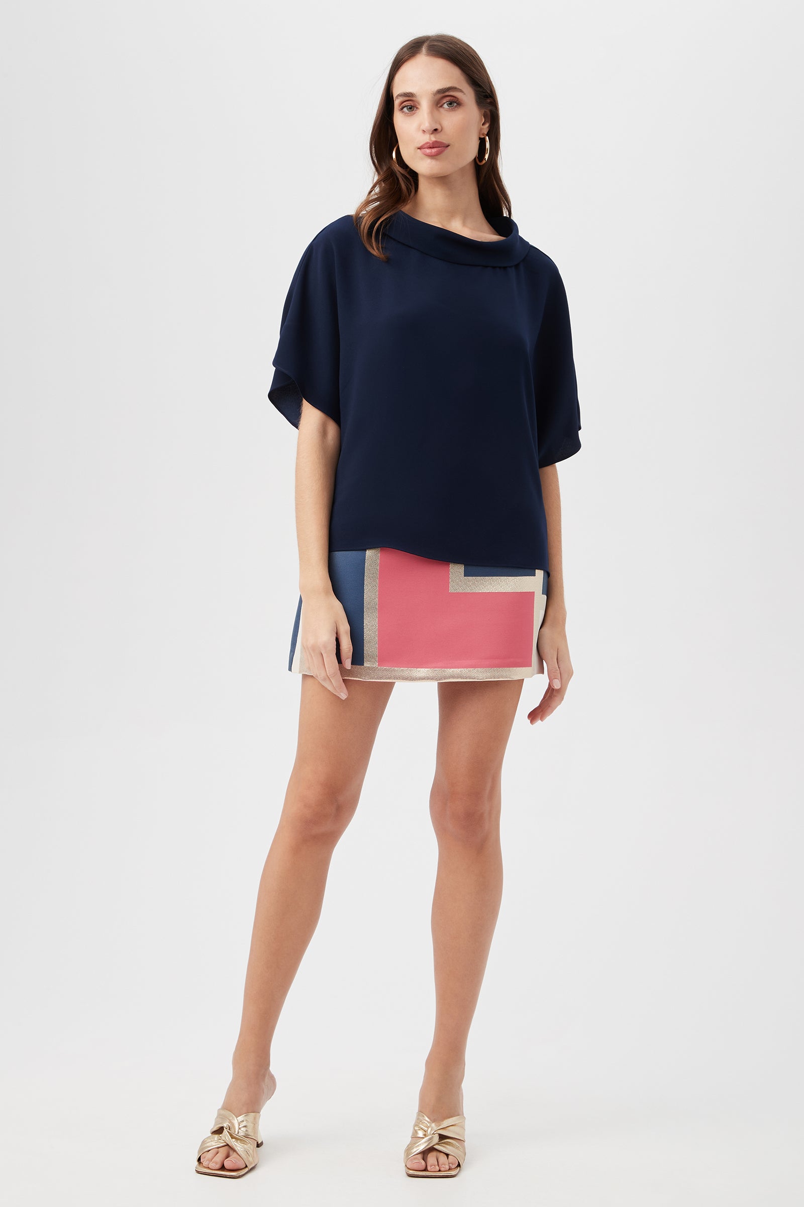 A woman with long brown hair and a neutral expression stands against a plain background, wearing the navy Honeysuckle Boatneck Dolman-Sleeve Top, a colorful geometric mini skirt in pink, blue, and beige, and gold sandals.