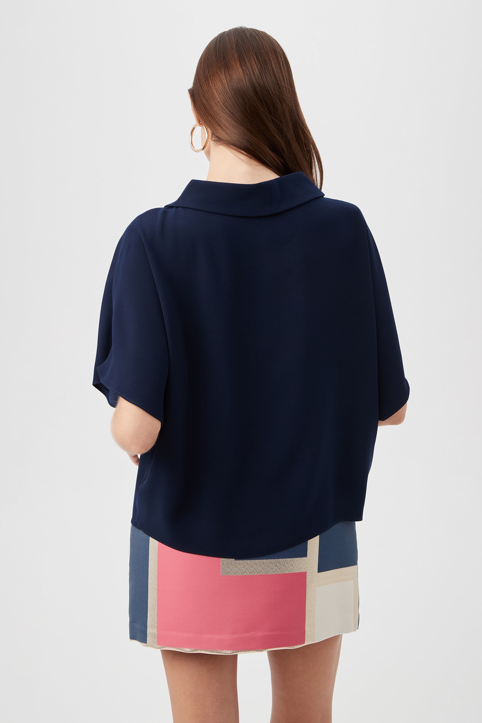 A woman with long brown hair, gold hoop earrings, and a navy Honeysuckle Boatneck Dolman-Sleeve Top paired with a pink, white, and blue color-block mini skirt is shown from the back against a plain backdrop.