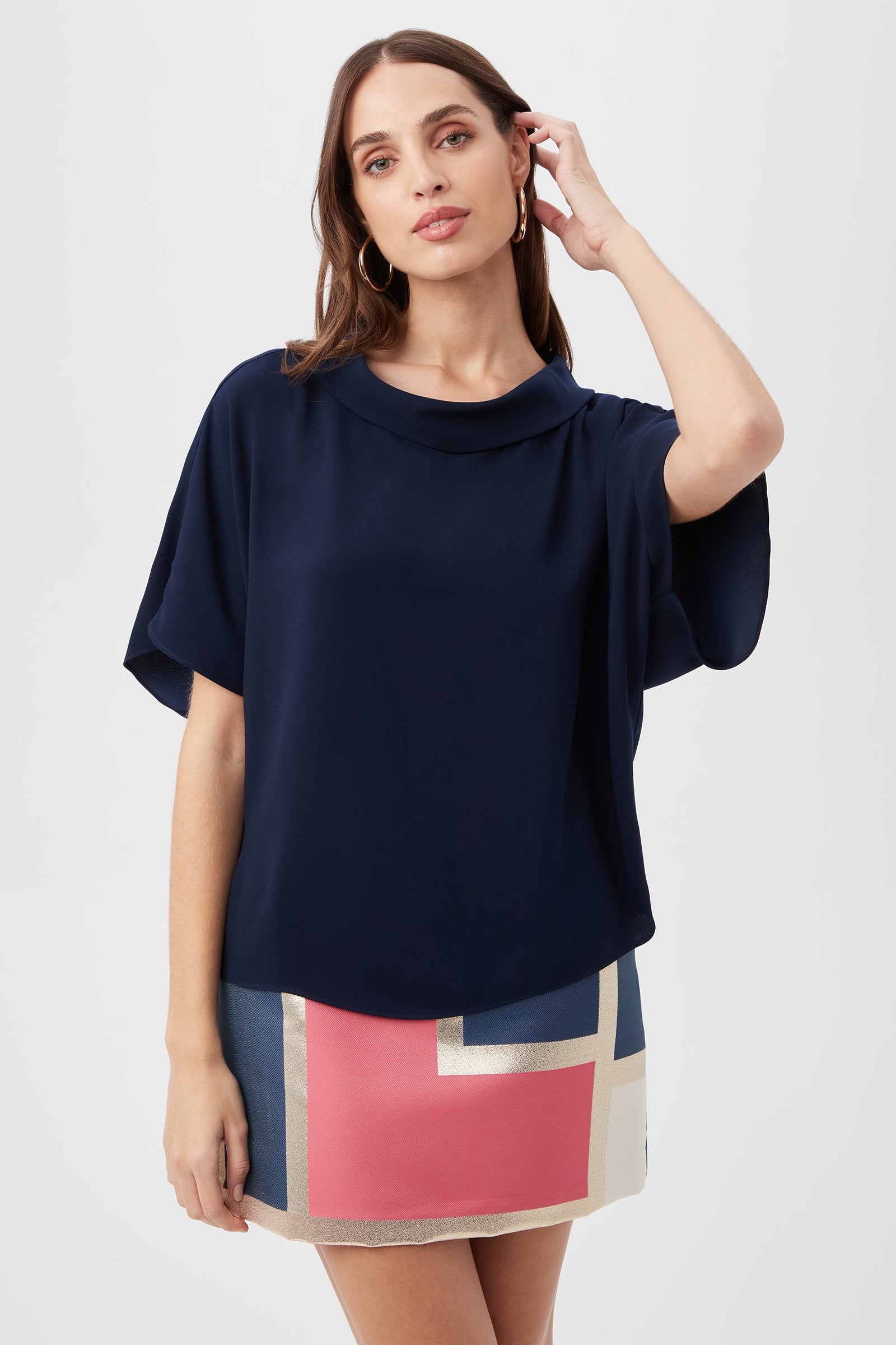 A woman with long brown hair wears the indigo Honeysuckle Boatneck Dolman-Sleeve Top and a mini skirt with bold pink, blue, and cream geometric blocks. She poses against a plain light background, touching her hair with one hand.