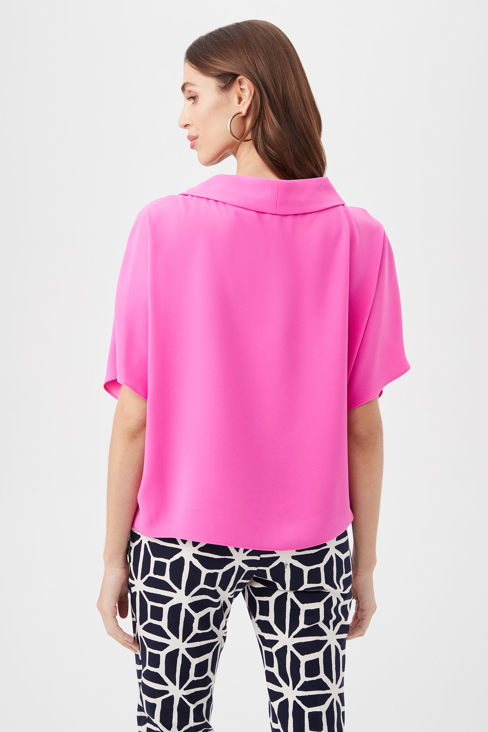 A woman with long brown hair and large hoop earrings faces away, wearing the Honeysuckle Boatneck Dolman-Sleeve Top in bright pink and black-and-white geometric patterned pants.