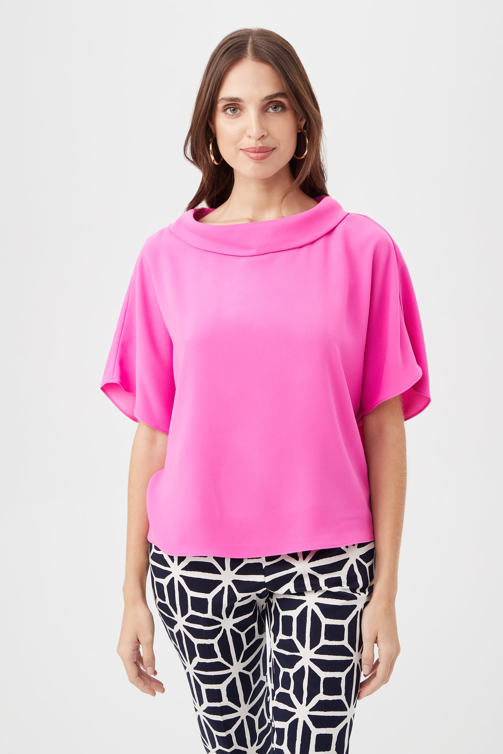 A woman wears the Honeysuckle Boatneck Dolman-Sleeve Top in Snapdragon, paired with black-and-white geometric patterned pants, standing facing forward against a plain white background.