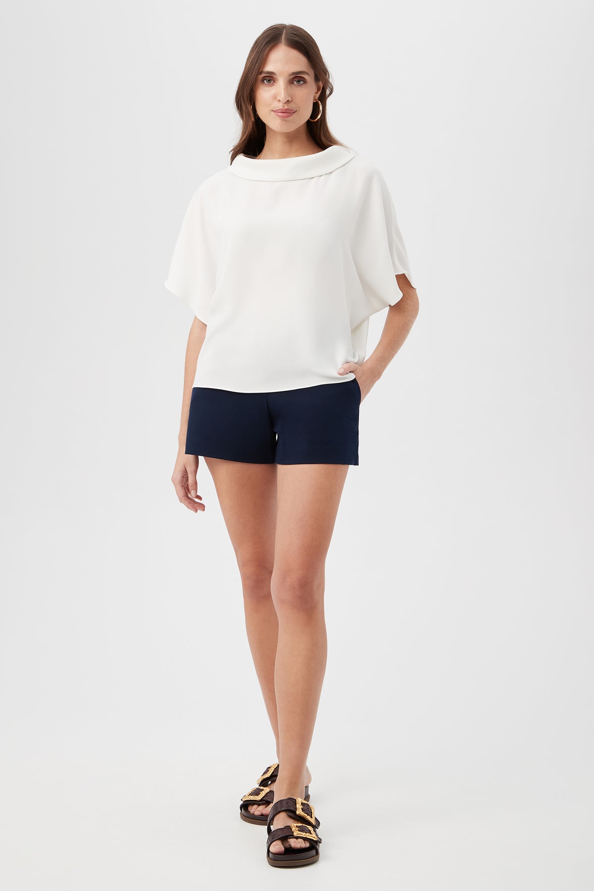 A woman stands against a plain white background wearing the Honeysuckle Boatneck Dolman-Sleeve Top, navy blue shorts, and black sandals with gold buckles. She has long brown hair and looks ahead with one hand in her pocket.