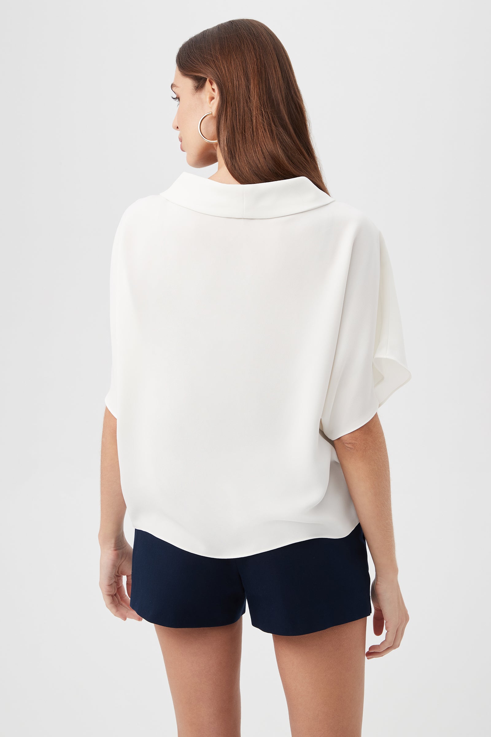 A woman with long brown hair, wearing the Honeysuckle Boatneck Dolman-Sleeve Top and navy blue shorts, stands facing away from the camera against a plain light background.