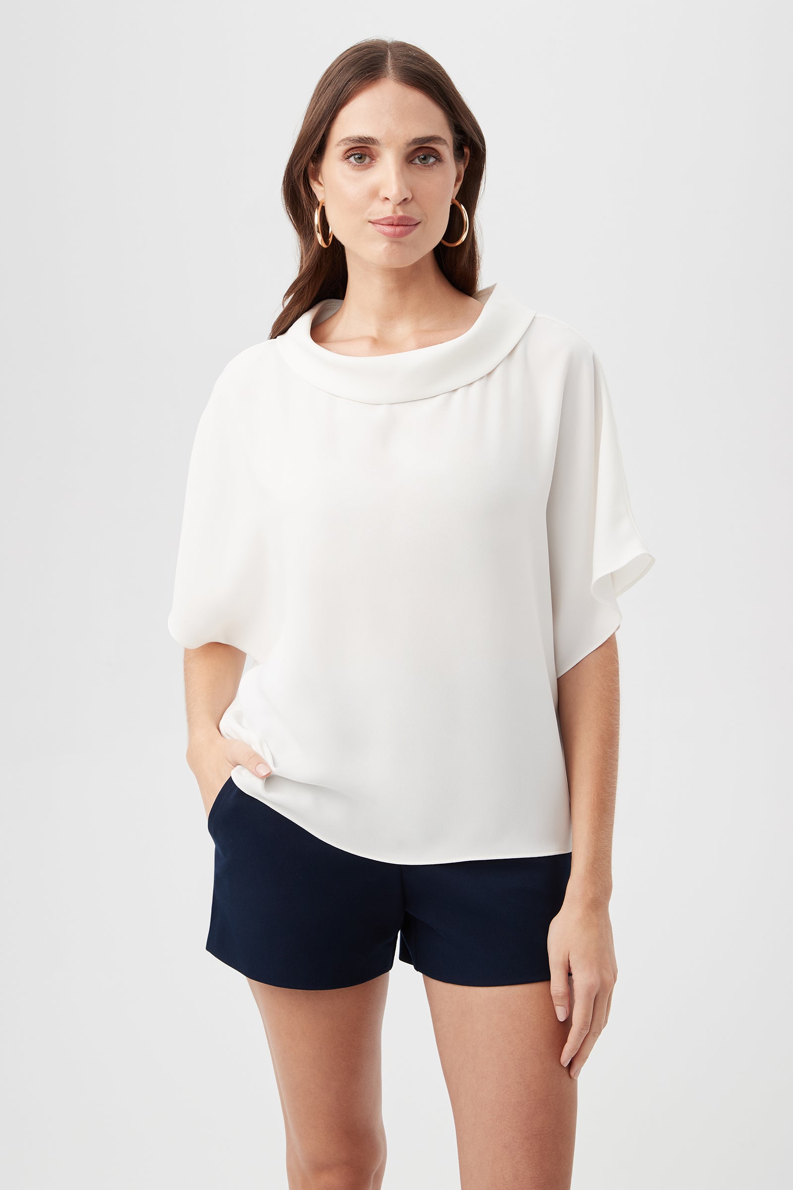 A woman with long brown hair and gold hoop earrings stands against a plain background, wearing the Honeysuckle Boatneck Dolman-Sleeve Top in Whitewash and black shorts, her expression neutral.