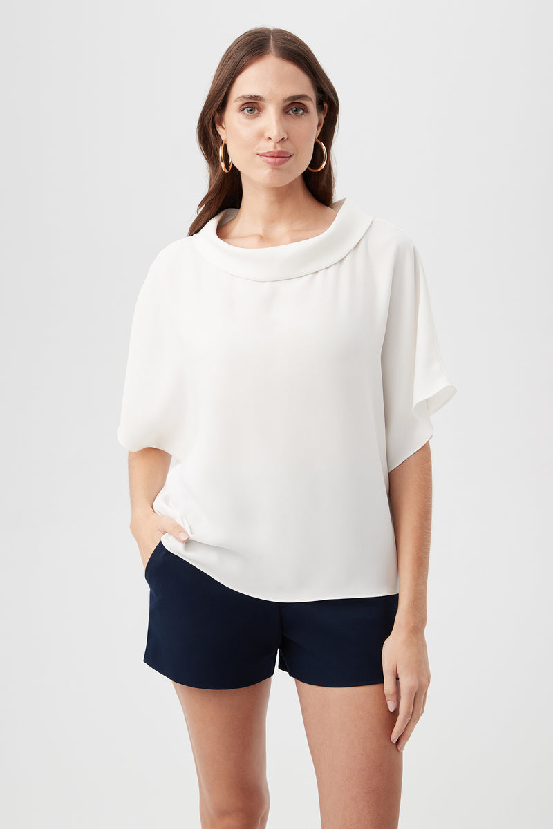 A woman with long brown hair and gold hoop earrings stands against a plain background, wearing the Honeysuckle Boatneck Dolman-Sleeve Top in Whitewash and black shorts, her expression neutral.