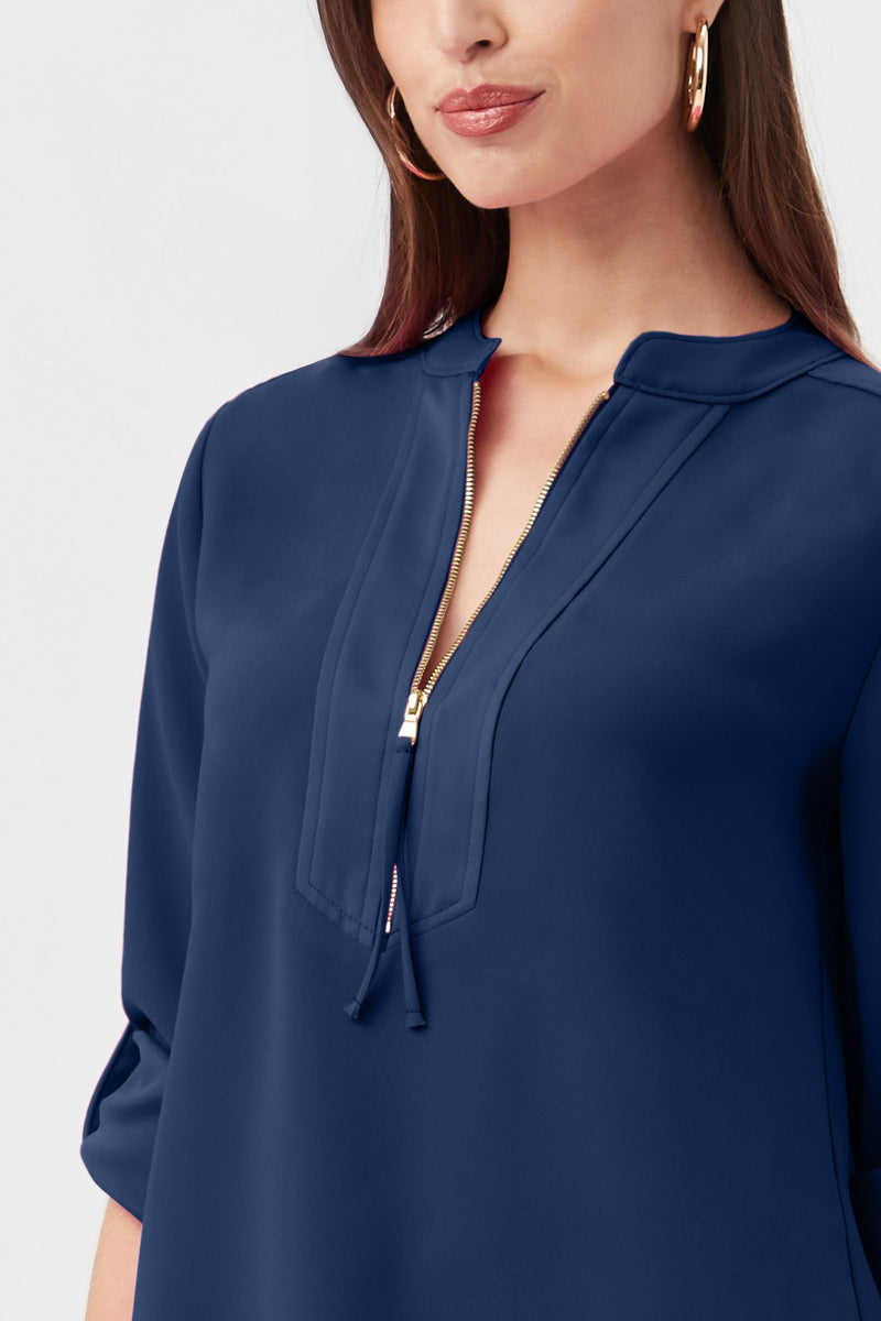 A woman with long brown hair and large hoop earrings is wearing the Kaiko 3/4-Sleeve Half-Zip Top, a navy blue blouse featuring a gold zipper at the collar and rolled-up sleeves. The image is cropped below her shoulders.