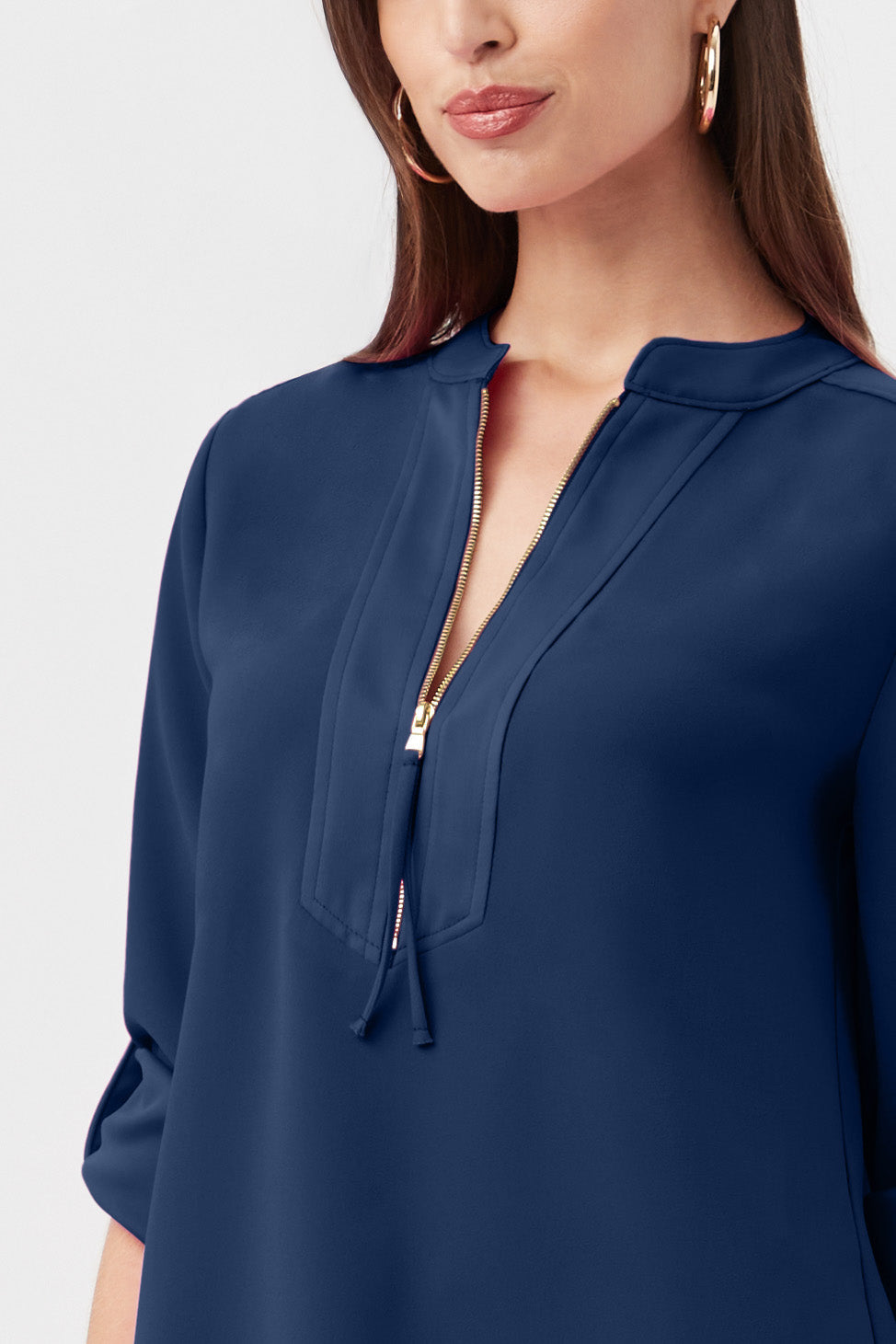 A woman with long brown hair and large hoop earrings is wearing the Kaiko 3/4-Sleeve Half-Zip Top, a navy blue blouse featuring a gold zipper at the collar and rolled-up sleeves. The image is cropped below her shoulders.