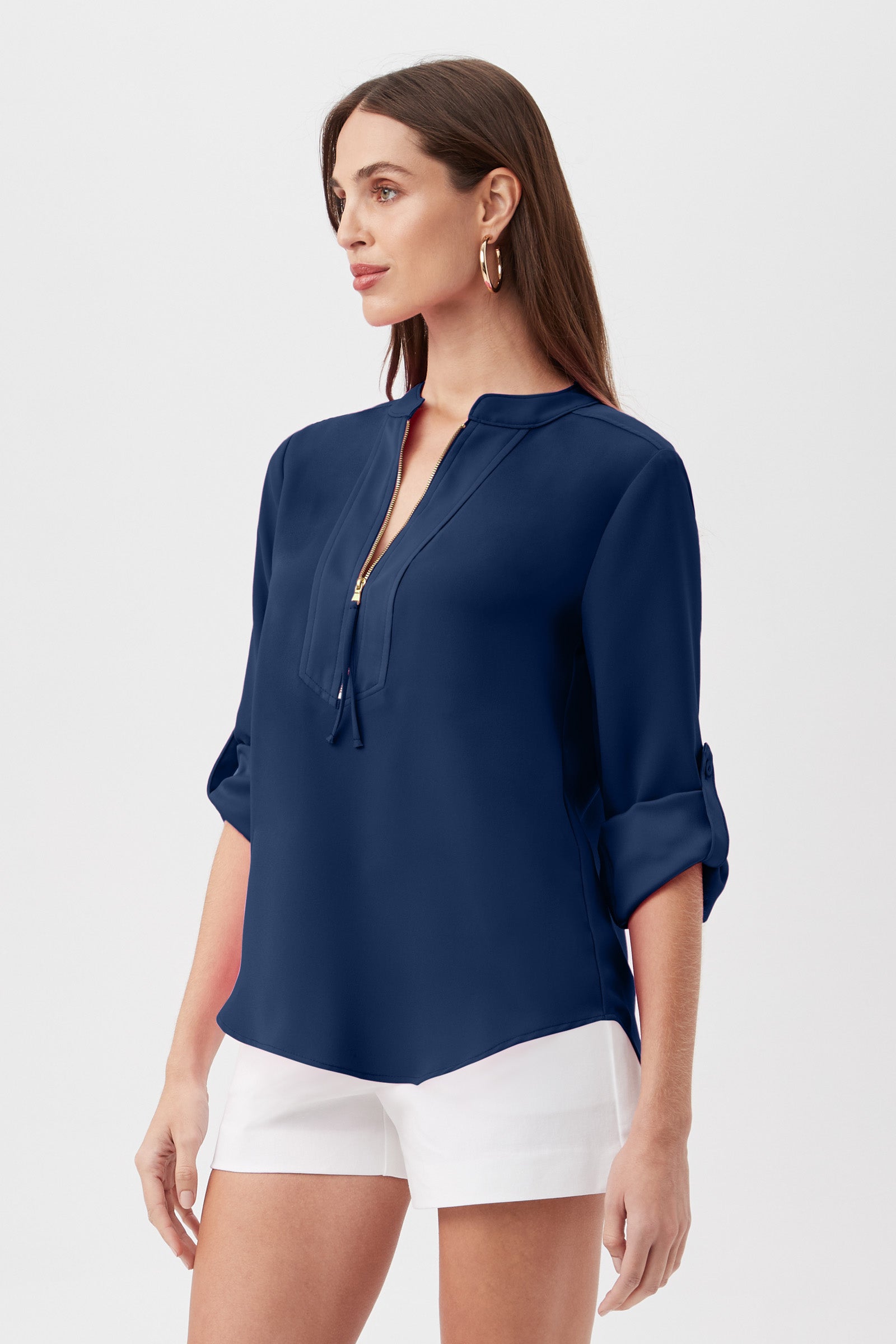 A woman with long brown hair wears the Kaiko 3/4-Sleeve Half-Zip Top in navy, featuring rolled-up sleeves, paired with white shorts. She stands against a plain light background, looking slightly to the side.