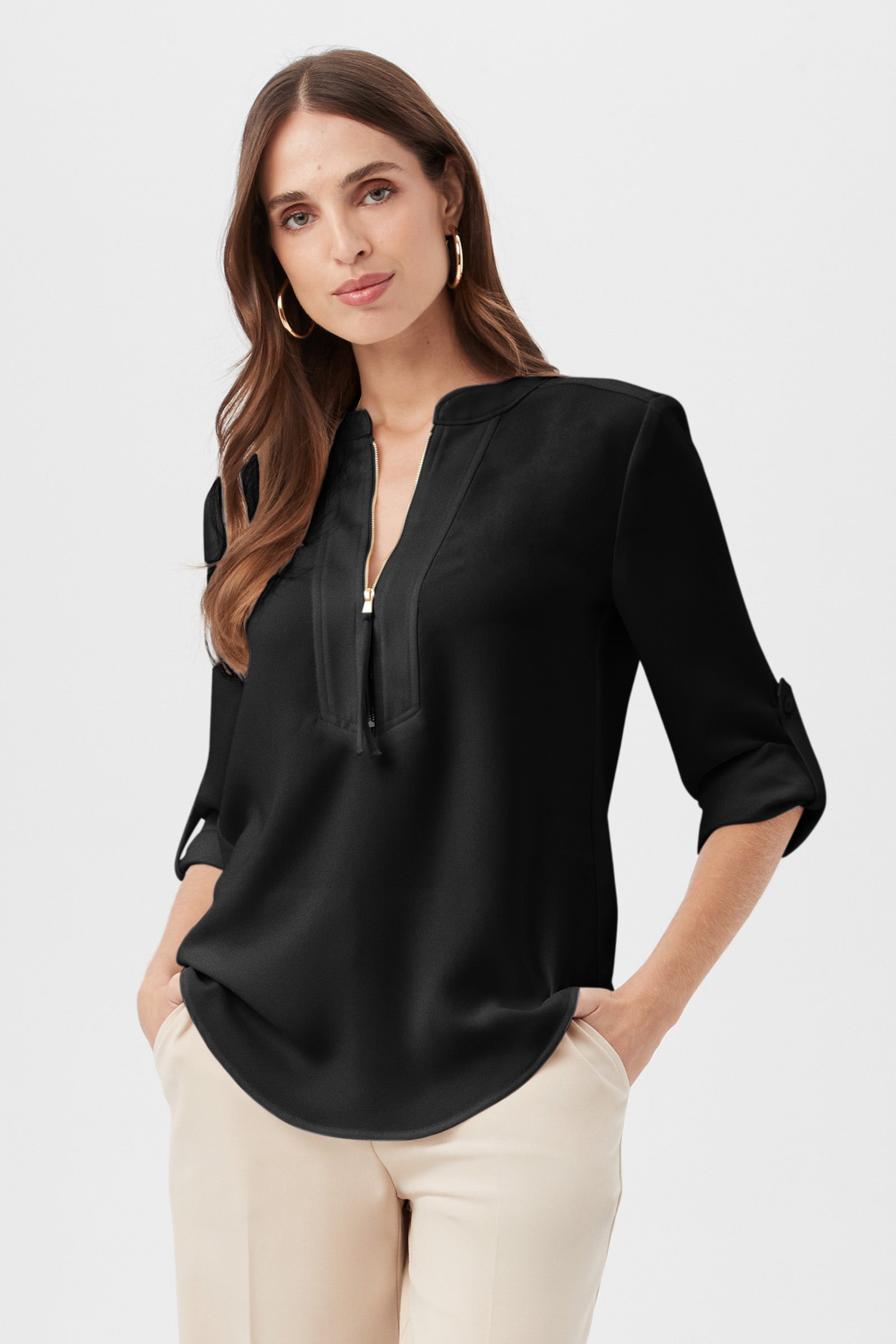 A woman wears the Kaiko 3/4-Sleeve Half-Zip Top in black, featuring a zippered V-neck and roll-tab sleeves, paired with light-colored pants. She stands with her hands in her pockets against a plain background.