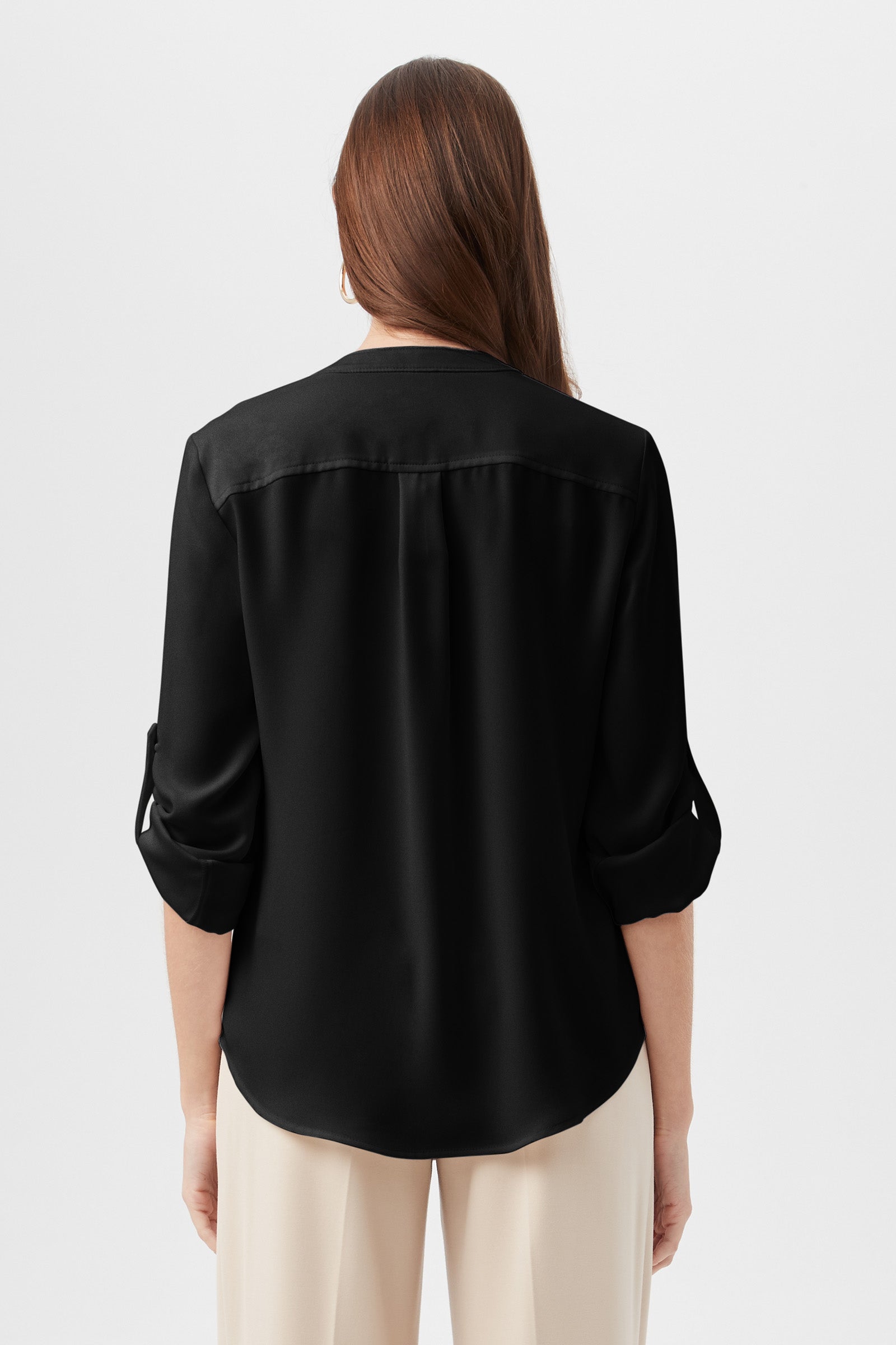 A woman with long brown hair is shown from the back, wearing a loose-fit black Kaiko 3/4-Sleeve Half-Zip Top and light-colored pants, standing against a plain white background.