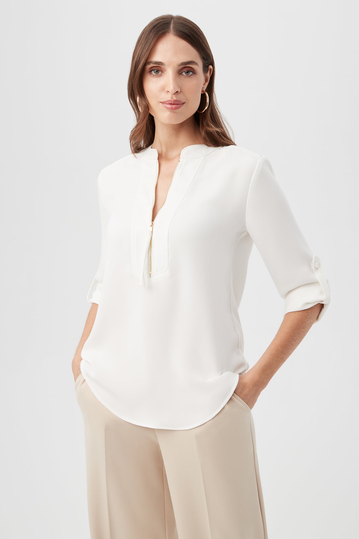 A woman with long brown hair wears the Kaiko 3/4-Sleeve Half-Zip Top in Whitewash and beige pants. She stands with hands in her pockets against a plain light backdrop, looking at the camera and smiling softly.