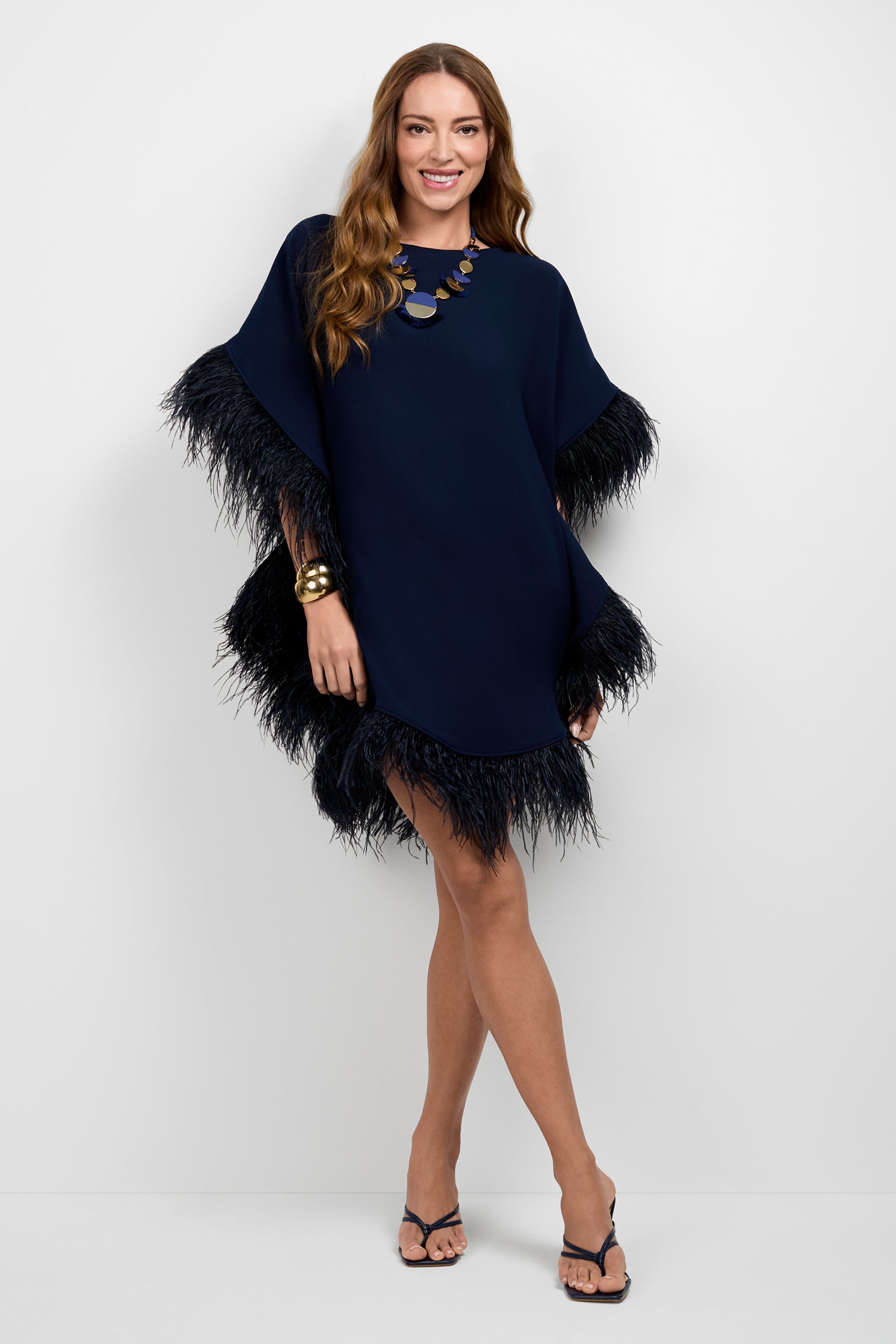 A woman stands against a white background, smiling with arms crossed, wearing the Mini Neena Caftan Dress in navy blue with feathered trim on the sleeves and hem, styled with a chunky necklace, bracelet, and black heeled sandals.
