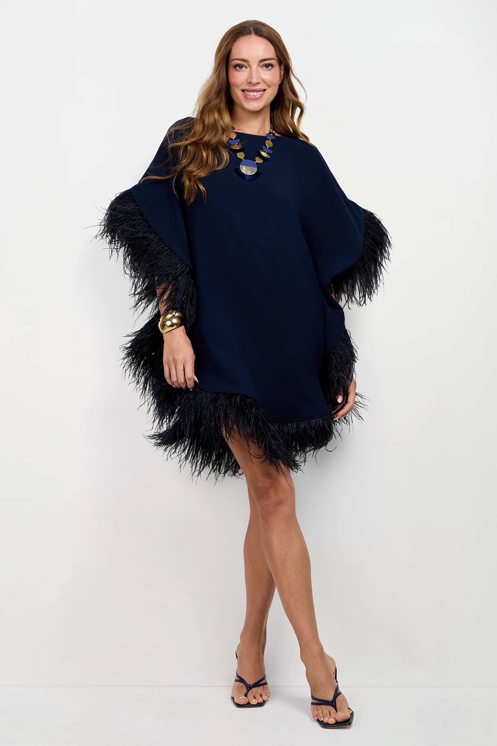A smiling woman poses against a white background in the Mini Neena Caftan Dress, a loose-fitting navy blue style with feathered trim on the sleeves and hem, paired with a chunky necklace, gold bracelet, and black heeled sandals.