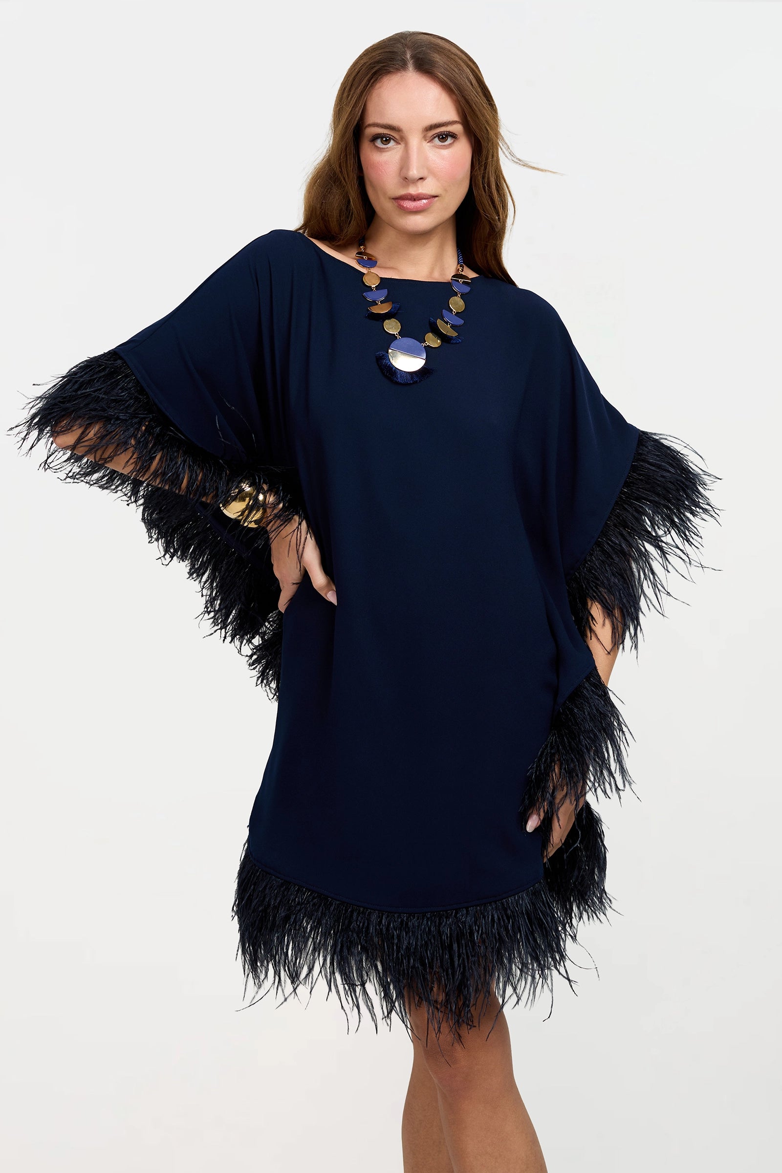 A woman models the Mini Neena Caftan Dress, a loose-fitting navy piece with black feather trim on the sleeves and hem. She accessorizes with a statement necklace and bracelet, standing against a plain white background.