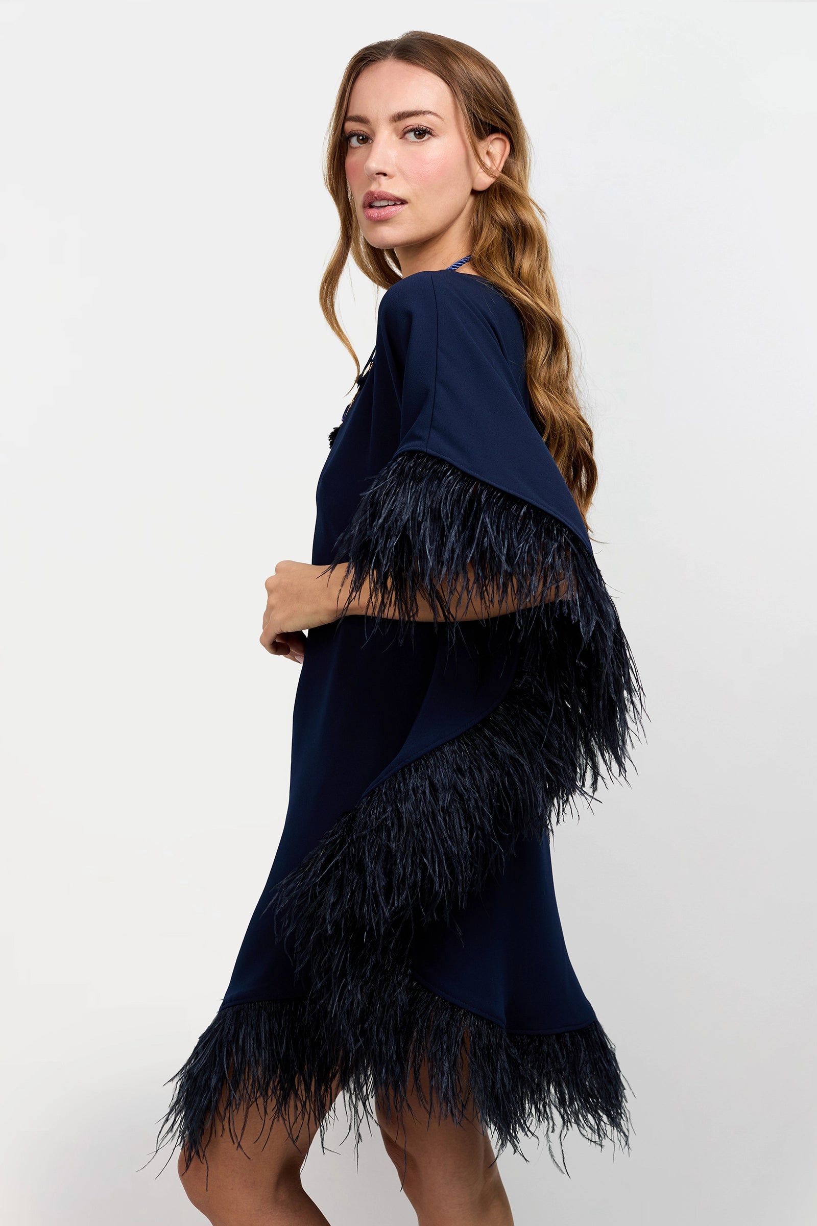 A woman with long light brown hair wears the Mini Neena Caftan Dress in navy blue with black feather trim on the sleeves and hem, standing against a plain light background and looking over her shoulder.