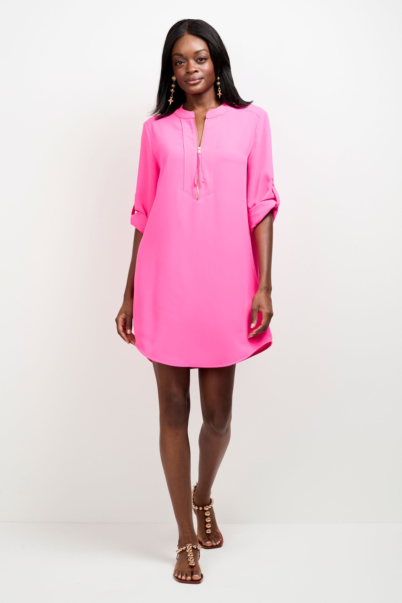 A woman stands against a white background wearing the Kaiko 2 Dress—a bright pink, long-sleeve, V-neck tunic with rolled-up sleeves—and brown studded sandals.