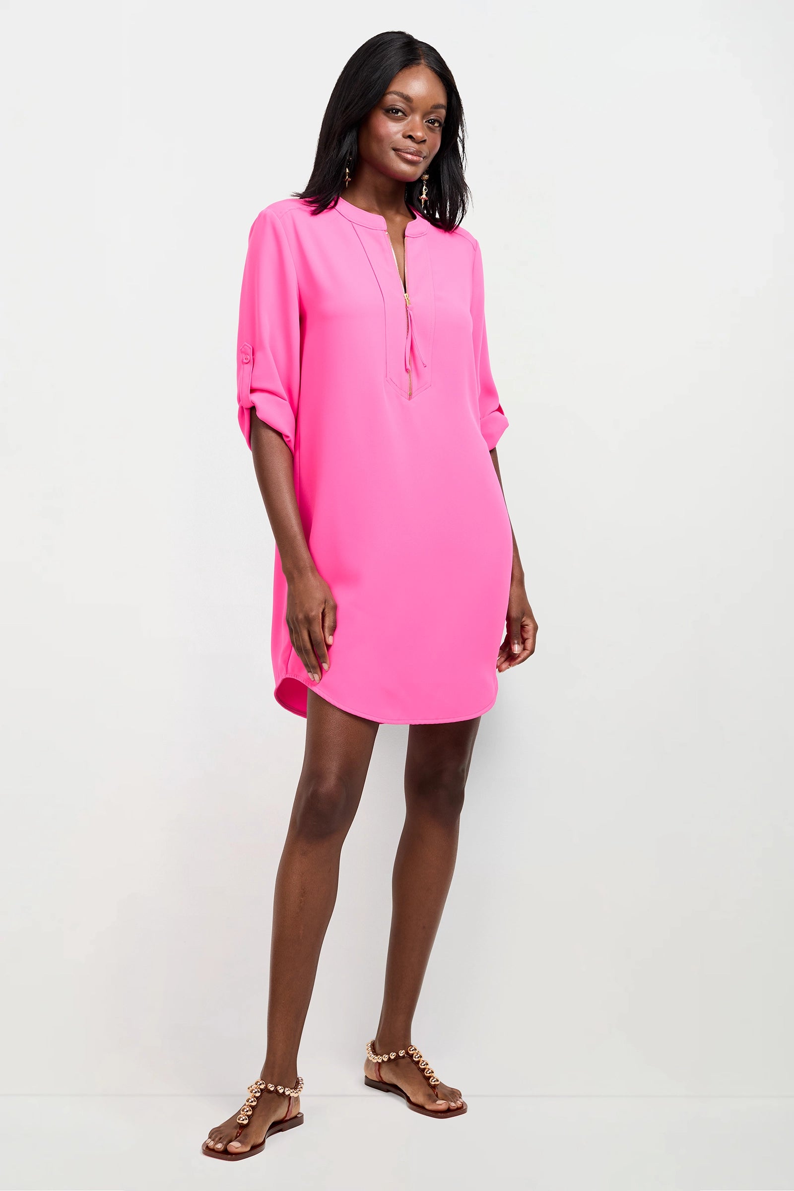 A woman stands against a plain white background wearing the Kaiko 2 Dress, a bright pink, long-sleeve, knee-length shift dress with a V-neck and roll-tab sleeves. She pairs it with brown flat sandals.