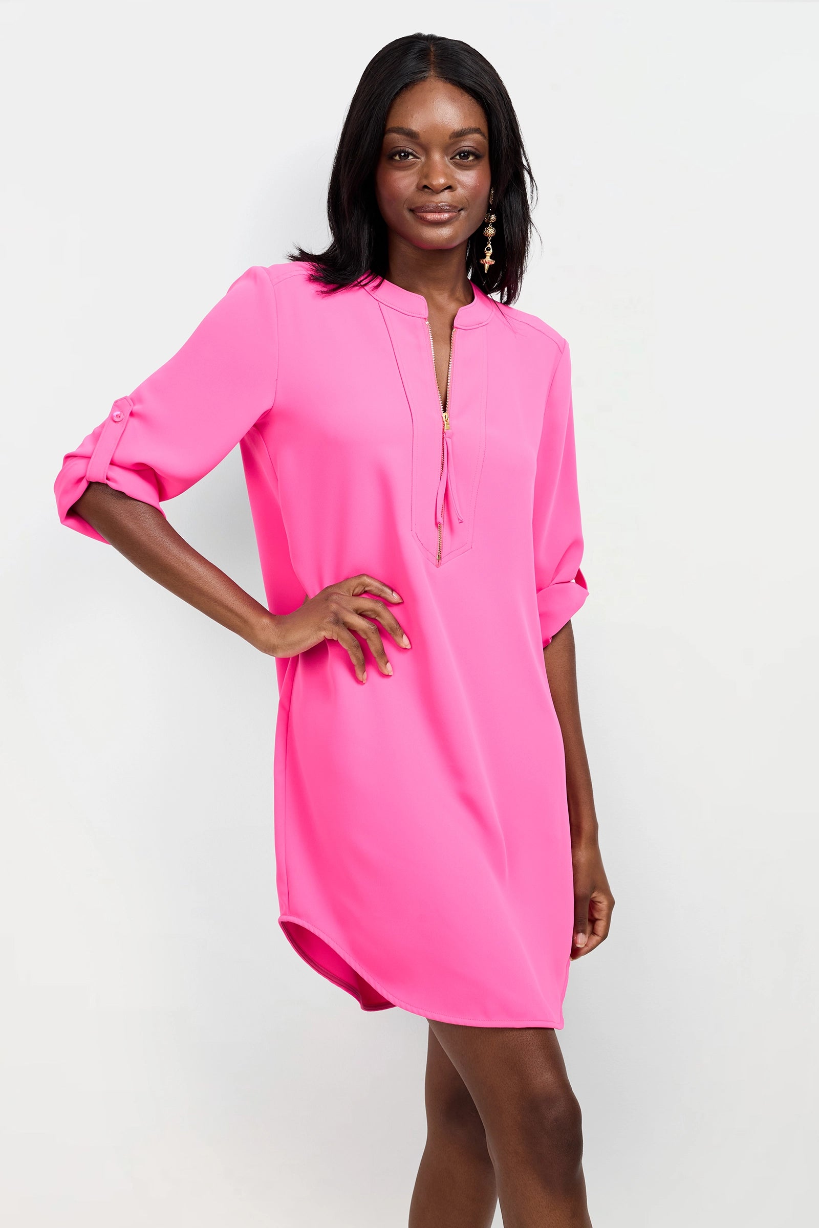 A woman smiles against a white background, wearing the Kaiko 2 Dress—a bright pink, knee-length dress with rolled-up sleeves and a V-neck—resting one hand on her hip.