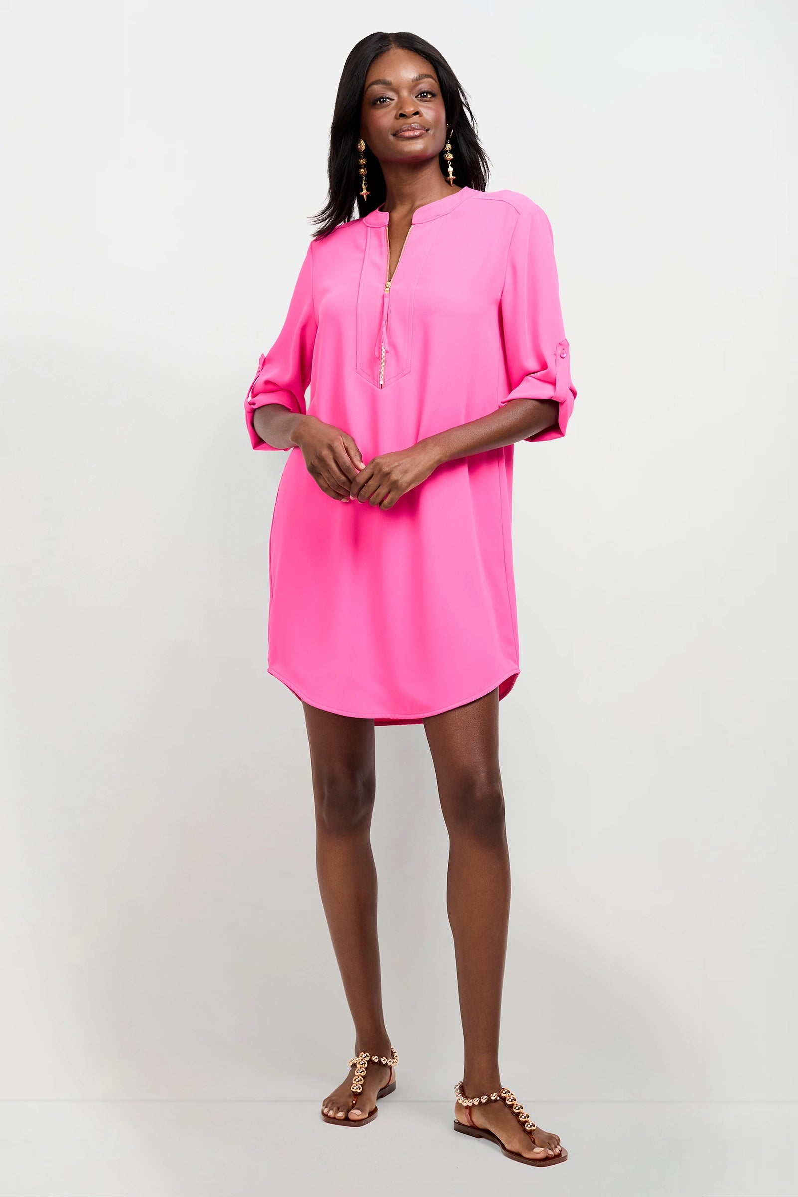 A woman smiles at the camera, wearing the Kaiko 2 Dress—a bright pink, loose-fitting tunic with rolled-up long sleeves—paired with gold earrings and brown flat sandals against a plain white background.