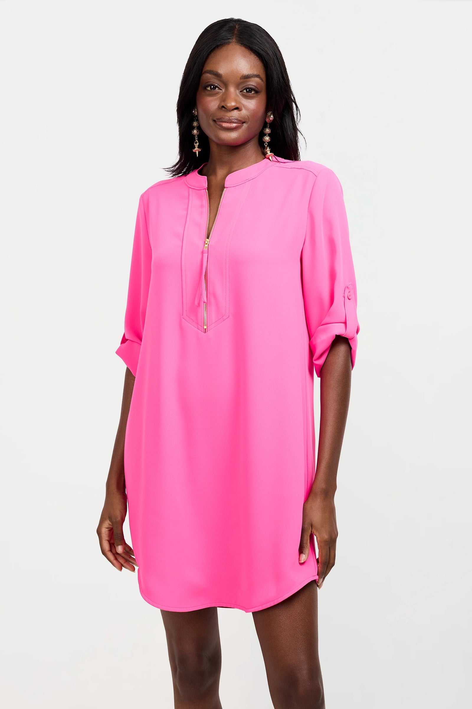 A woman with long dark hair and dangling earrings stands against a plain background, smiling and wearing the Kaiko 2 Dress—a bright pink, long-sleeve, loose-fitting dress with a partial zip-up front.