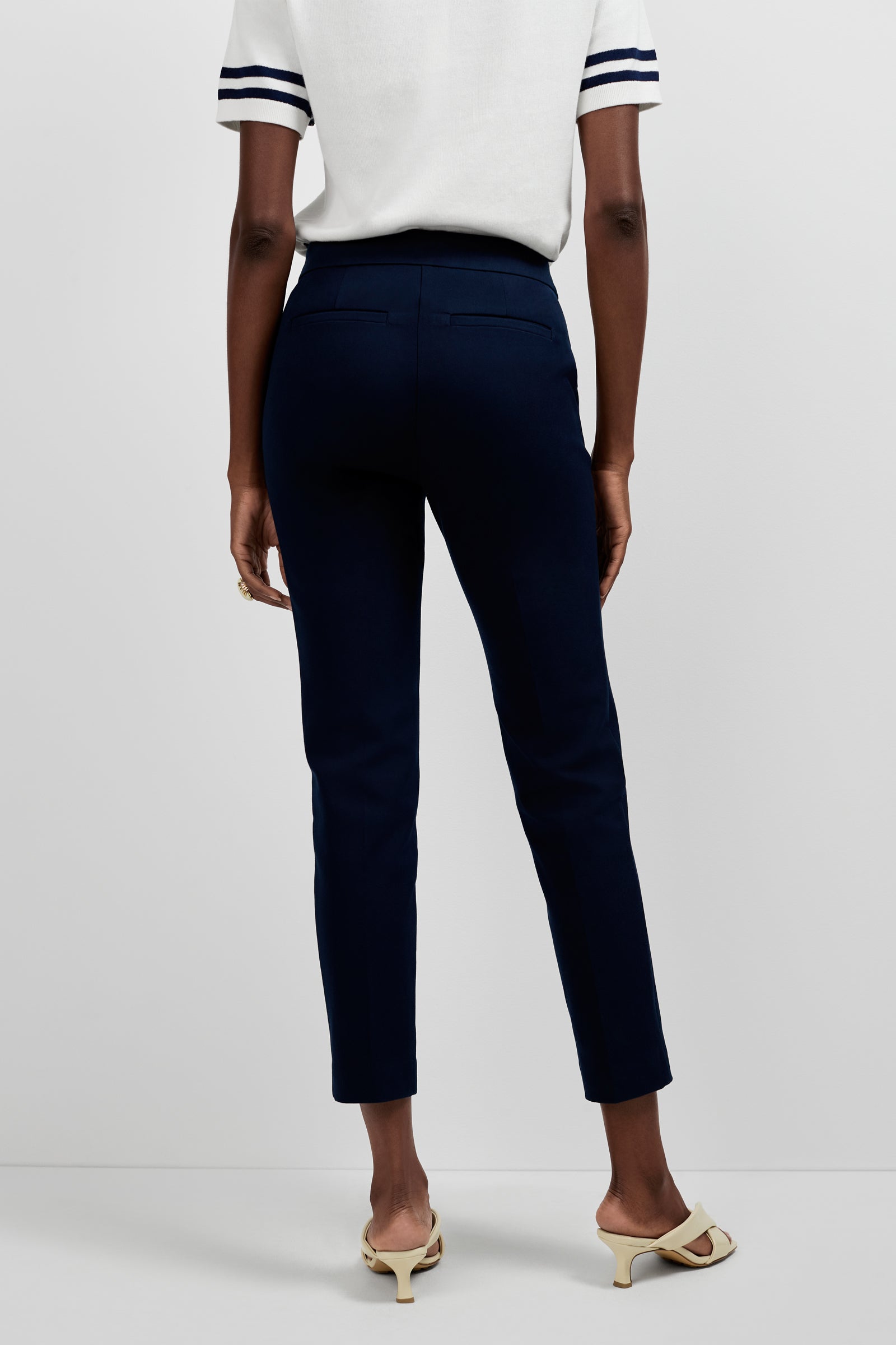 A person stands facing away, wearing Aubree 2 Cigarette Pants in dark navy, a short-sleeve white top with black-striped sleeves, and light-colored heeled sandals against a plain background.