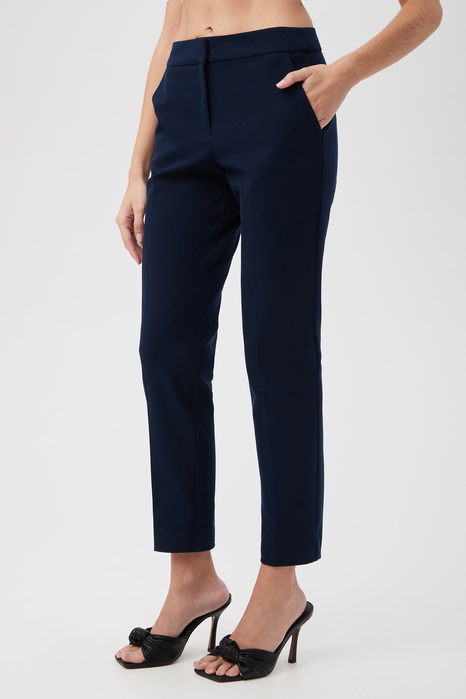 A person stands against a plain light background wearing Aubree 2 Cigarette Pants in navy blue and black high-heeled sandals, with one hand in their pocket.