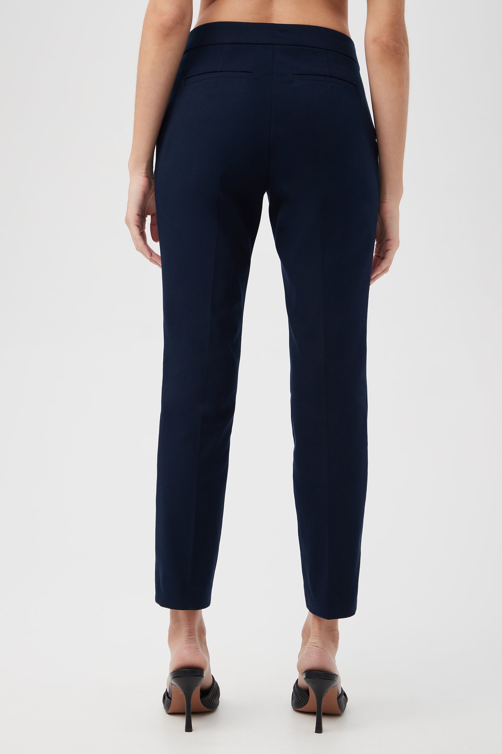 A woman stands facing away, wearing Aubree 2 Cigarette Pants in navy blue with black high-heeled shoes against a plain white background.