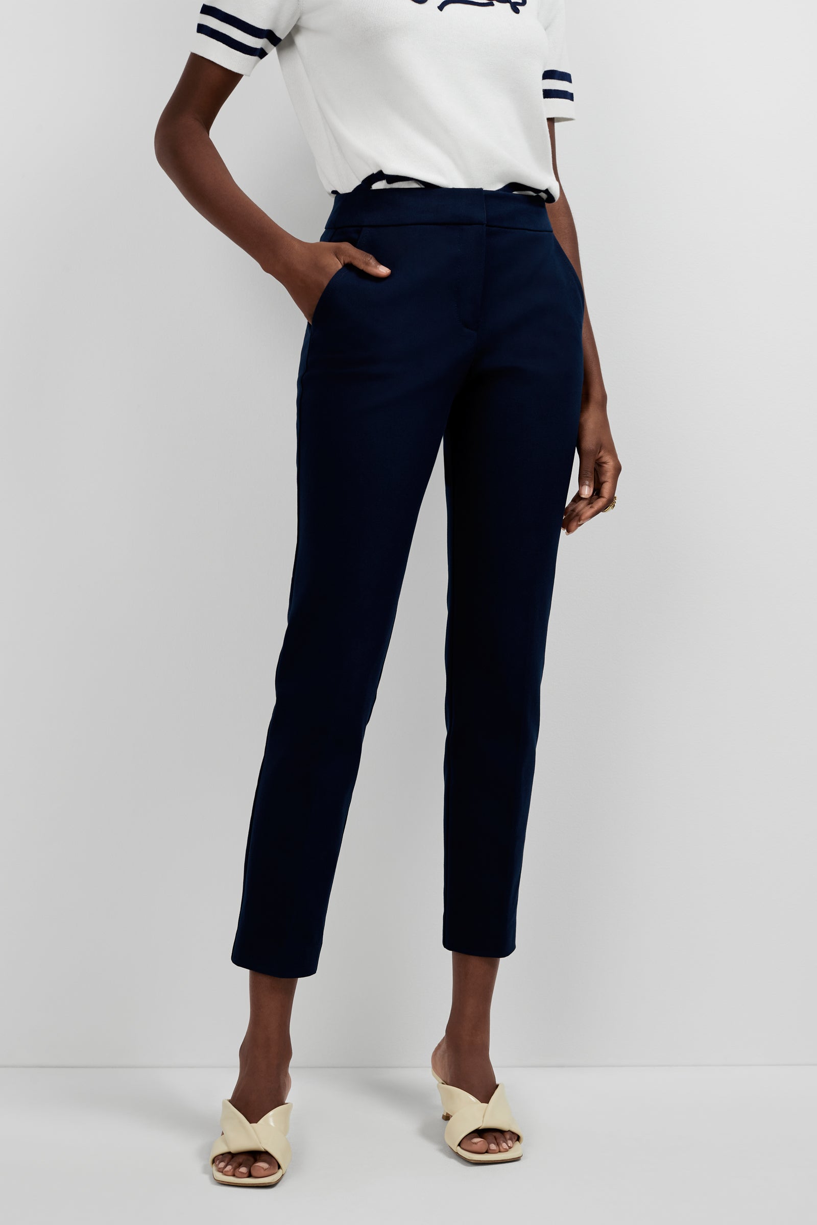 A person poses against a plain background, wearing a white short-sleeved top with dark stripes, cream open-toed heeled sandals, and the Aubree 2 Cigarette Pants in navy blue high-waisted ankle style, with one hand in their pocket.